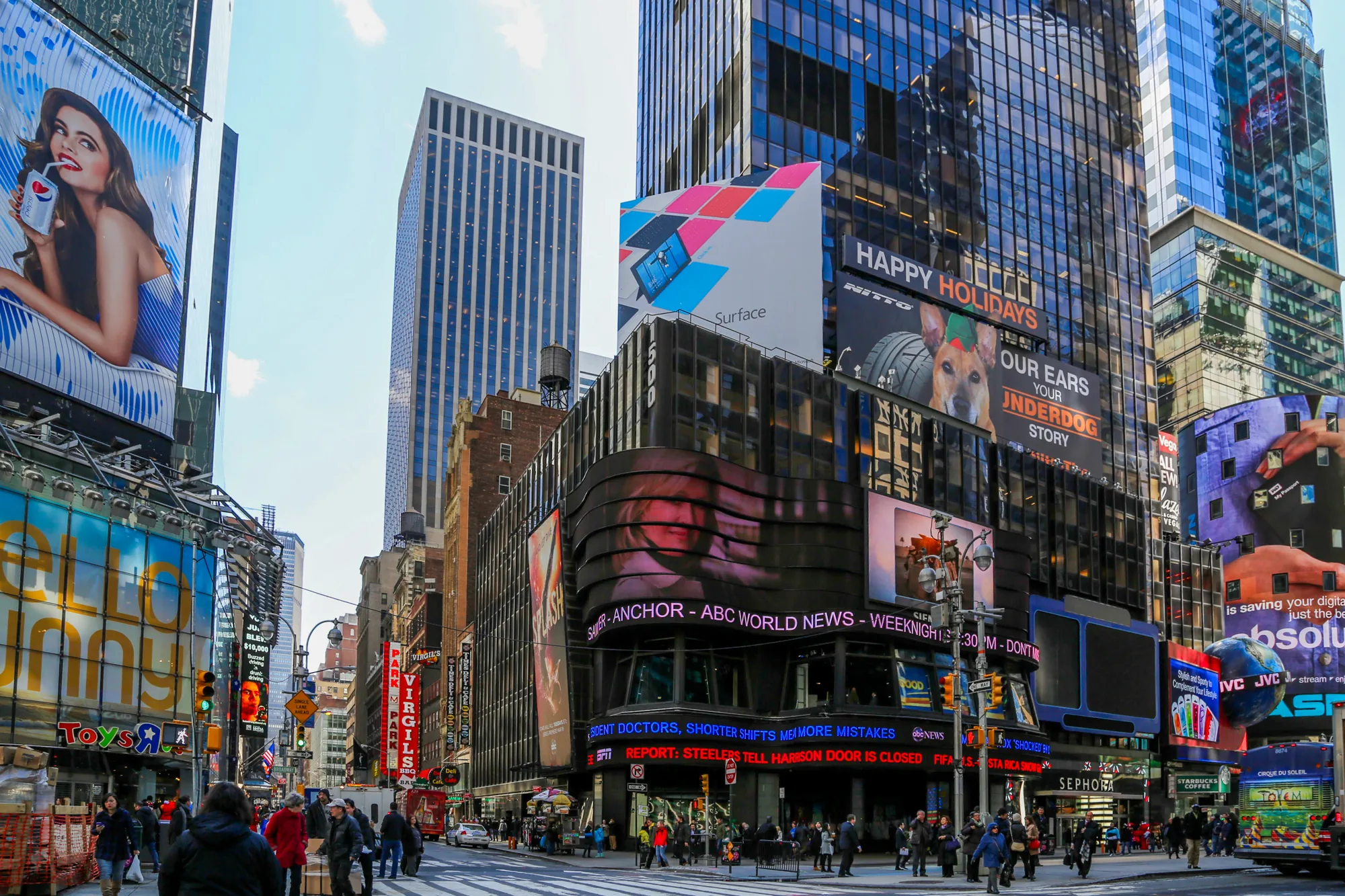 この写真は、アメリカ合衆国ニューヨーク市の賑やかな街並みを捉えたものです。高層ビルが林立し、その壁面には巨大な広告が所狭しと掲げられています。ペプシ、マイクロソフトのSurface、NITTOタイヤ、スターバックス、TOYS”R”USなどの広告が見られます。ABCニュースの広告や、スポーツニュースの告知も掲示されています。街には多くの人が行き交い、活気に満ち溢れています。信号機や街灯が設置され、都会のインフラが整備されている様子が伺えます。全体的に明るい時間帯に撮影された写真で、ニューヨークのエネルギーと多様性を感じさせる一枚です。