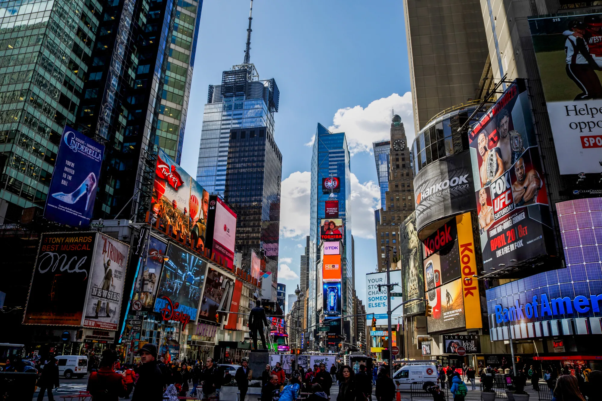 この写真は、アメリカ合衆国ニューヨークのタイムズスクエアで撮影されたものです。画面全体に高層ビルが立ち並び、その壁面には巨大な広告看板が所狭しと掲げられています。これらの広告は、ミュージカル（シンデレラ、ワンスなど）、企業（コダック、バンクオブアメリカ、TDK、東芝、LG）、スポーツイベント（UFC）など多岐にわたります。  通りは多くの人々で賑わっており、様々な人種や服装の人々が歩いています。彫刻が通りの中央に配置されており、観光客や地元の人々がその周りに集まっています。画面上部には青空が見え、ビル群の隙間から光が差し込んでいます。全体的に、活気に満ちた都会の雰囲気と、観光地としての賑わいが伝わってきます。色彩豊かでエネルギーに満ちた写真です。