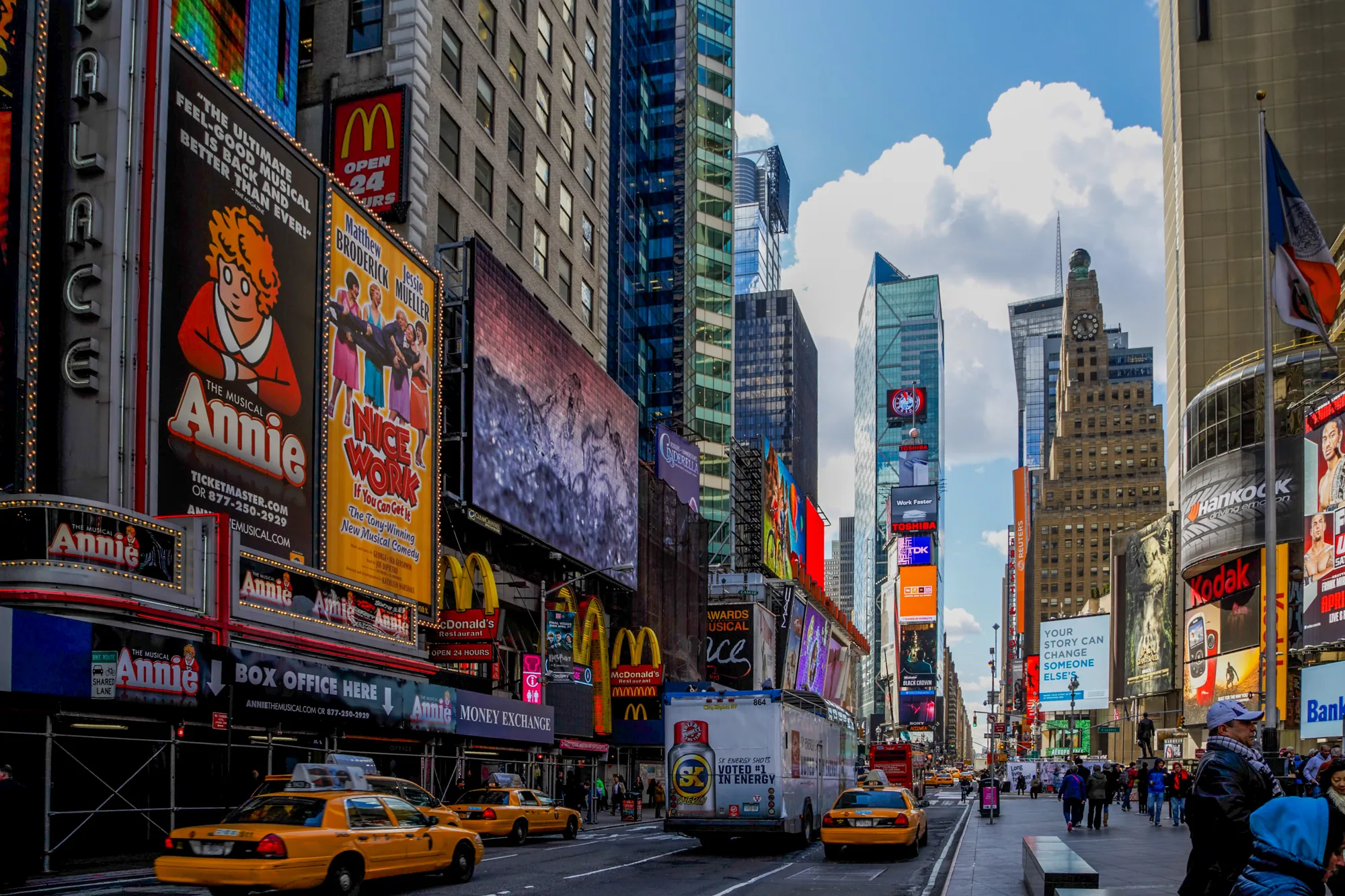 この写真はアメリカ合衆国ニューヨーク市のタイムズスクエアで撮影されたものです。高層ビルが立ち並び、巨大な広告看板がひしめき合っています。ミュージカル「アニー」や「NICE WORK」の看板が目立つほか、マクドナルドやコダックなどの企業広告も確認できます。街には多くの人が行き交い、ニューヨーク名物の黄色いタクシーが何台も走っています。空は青く、白い雲が浮かんでおり、活気のある都市の雰囲気を伝えています。全体的に明るく賑やかな印象を与える写真です。