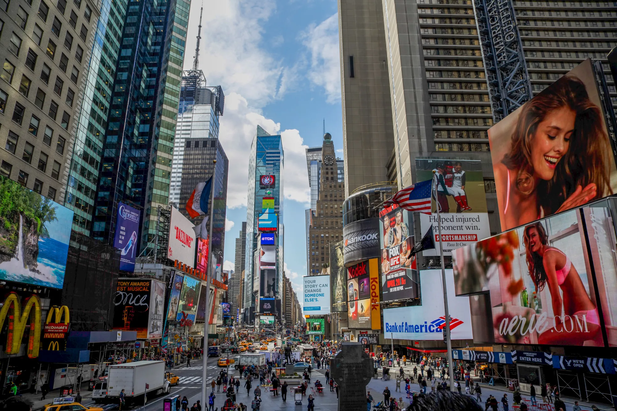写真はアメリカ合衆国ニューヨークのタイムズスクエアを捉えたものです。高層ビルが立ち並び、その壁面には巨大な広告看板が数多く掲げられています。マクドナルドの「M」のロゴや、ミュージカルのポスター、アパレルブランドの広告など、様々な種類の広告がひしめき合っています。道路には多くの人々が行き交い、タクシーが走っており、活気に満ちた様子が伝わってきます。空には青空が広がり、一部に雲が見られます。全体的に、タイムズスクエアの喧騒と賑やかさを表現した写真です。