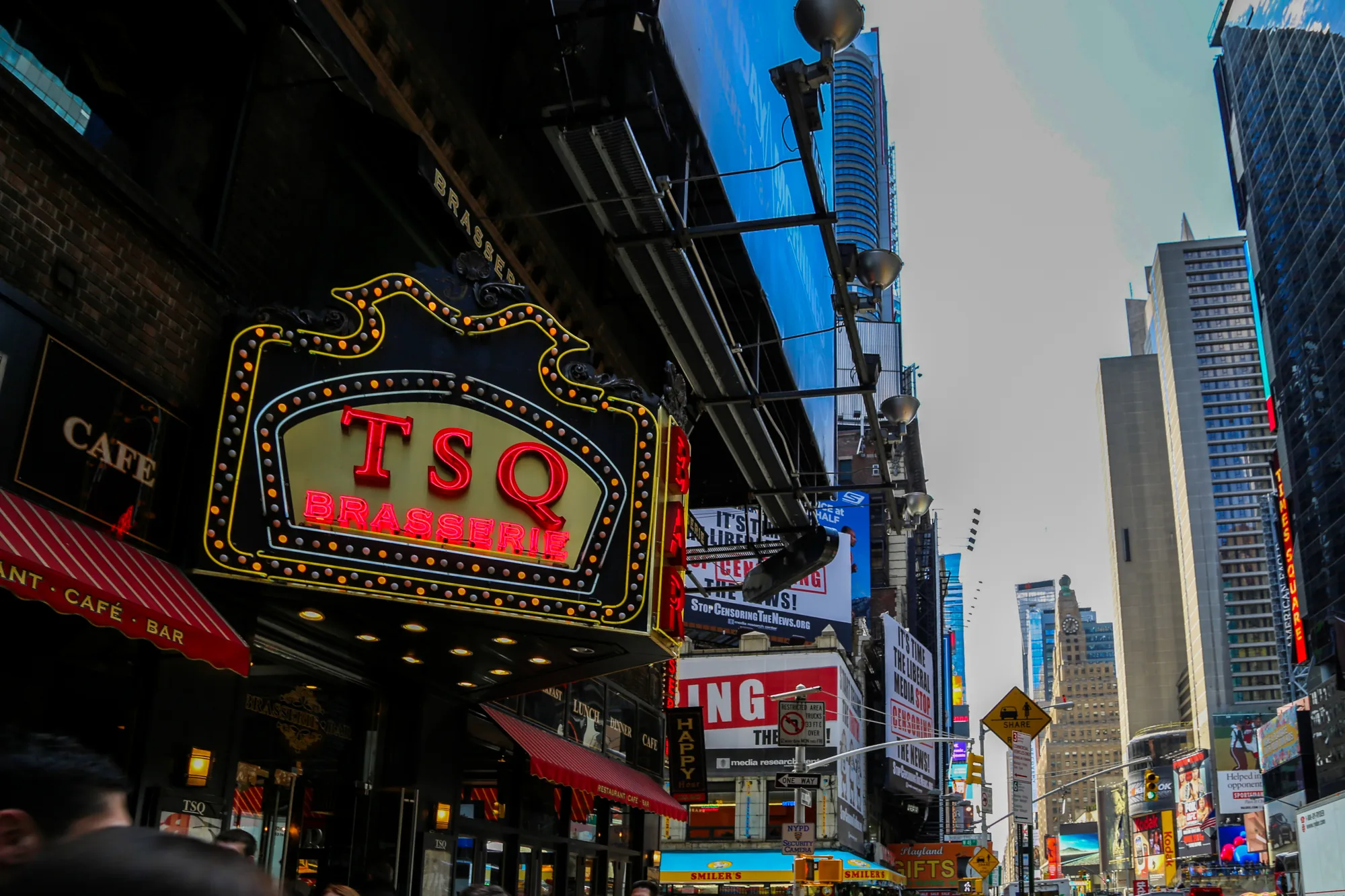 この写真は、アメリカ合衆国ニューヨークのタイムズスクエアで撮影されたものです。画面左側には、電飾で飾られた「TSQ BRASSERIE」というレストランの看板が大きく写っています。赤と黄色のネオンサインが目を引き、活気のある雰囲気を出しています。レストランの赤いストライプの庇も印象的です。  写真全体は、無数の看板や広告、そして高層ビル群で埋め尽くされています。雑多な看板の数々は、タイムズスクエアならではの喧騒とエネルギーを表現しています。空は薄曇りで、高層ビルの輪郭がぼんやりと浮かび上がっています。写真の下部には、人々の頭の一部が写っており、タイムズスクエアの賑わいを伝えています。全体的に、タイムズスクエアの象徴的な風景を捉えた一枚と言えるでしょう。