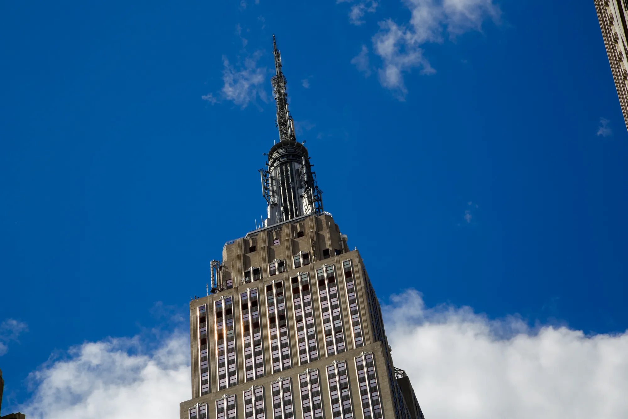 写真には、ニューヨークの象徴的な建造物であるエンパイアステートビルの上部が、見上げるようなアングルで捉えられています。建物はアールデコ調のデザインで、幾何学的な形状が特徴的です。窓が整然と並び、光を反射して輝いています。ビルの頂上には、アンテナがそびえ立っており、空に向かって伸びています。背景には、鮮やかな青空と白い雲が広がっており、建物を一層引き立てています。全体的に、力強く、象徴的な雰囲気が感じられます。