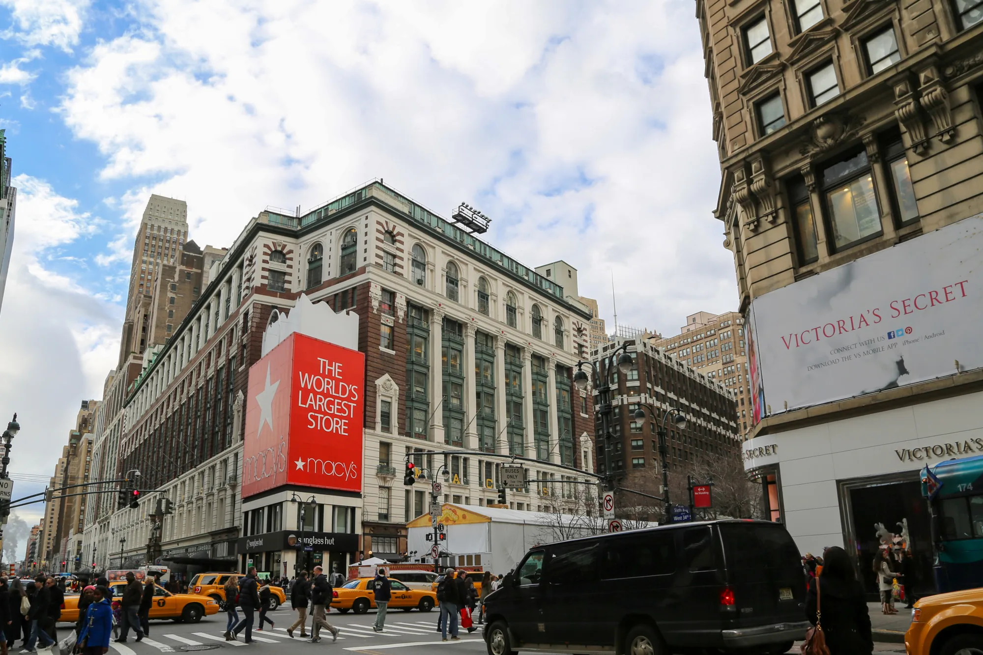 写真は、アメリカ合衆国のニューヨークの街並みを捉えたものです。画面中央には、巨大な「THE WORLD'S LARGEST STORE Macy's」という赤い看板が掲げられた大きな建物が写っています。この建物の隣には「VICTORIA'S SECRET」の大きな広告看板が見えます。  画面下部には、横断歩道を歩く人々や、ニューヨーク名物の黄色いタクシーが複数台写っており、街の賑わいを表現しています。画面奥には高層ビル群が立ち並び、ニューヨークらしい都会的な風景が広がっています。空は曇っており、全体的に落ち着いた雰囲気の写真です。