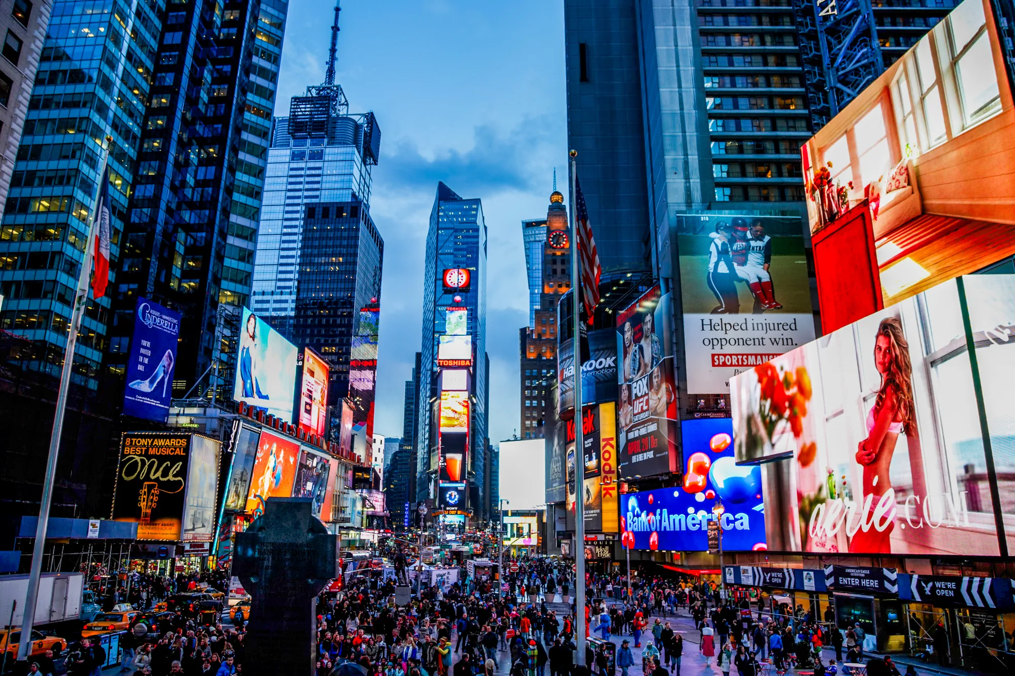 この写真は、アメリカ合衆国のニューヨークにあるタイムズスクエアで撮影されたものです。空は夕暮れ時の青色で、高層ビル群がそびえ立っています。ビルには巨大な広告がいくつも掲示されており、ネオンライトや鮮やかな色彩で埋め尽くされています。「Cinderella」「TOSHIBA」「Bank of America」などの広告が確認できます。  道路は人で埋め尽くされており、多くの人々が様々な方向へ歩いています。画面左には黄色のタクシーがいくつか見えます。タイムズスクエアの賑やかで活気のある雰囲気が伝わってきます。全体的に、都市のエネルギーと商業的な輝きを感じさせる写真です。