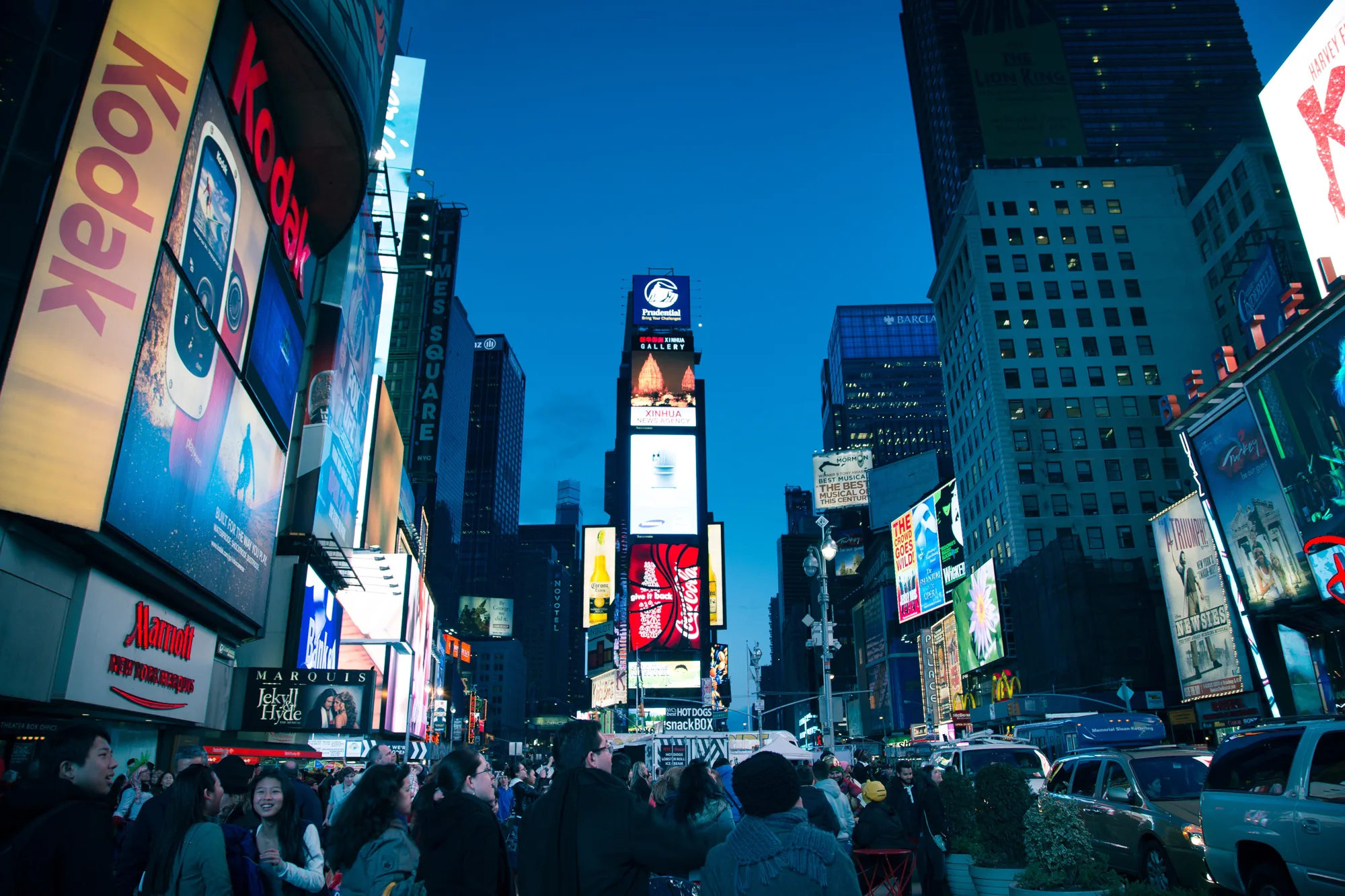 この写真は、アメリカ合衆国ニューヨークのタイムズスクエアで撮影されたものです。夕暮れ時か夜のようで、空は濃い青色をしています。画面全体に高層ビルが立ち並び、その壁面には巨大な広告看板が多数設置されています。KodakやMarriottの看板が見え、多くの観光客や通行人がそれらを眺めています。人々は冬の装いで、コートや帽子を着用しています。画面中央にはPrudentialのロゴが入った巨大なデジタルサイネージがあり、その下にはXinhua News Agencyやコカ・コーラなどの広告が表示されています。通りの向こうにはホットドッグの屋台も見えます。写真全体から、タイムズスクエアの活気と賑わいが伝わってきます。