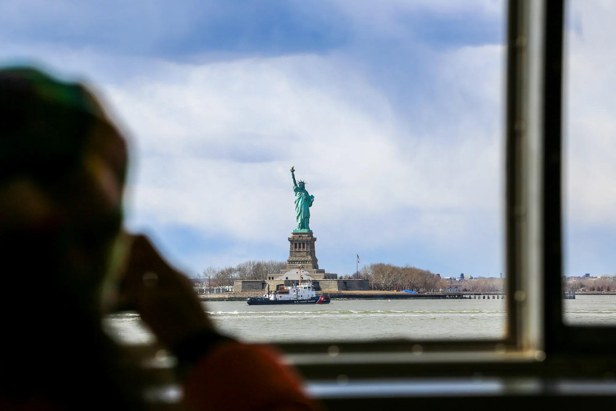 この写真は、アメリカ合衆国ニューヨークで撮影されたものです。窓枠を通して自由の女神像が見えます。視界には、自由の女神像の台座の下を航行する沿岸警備隊の船が写っています。前景には、おそらくフェリーや観光船の乗客のシルエットがぼんやりと写っており、窓から外を眺めている様子がうかがえます。背景には、水面と、うっすらと見える陸地が広がっています。空は晴れており、いくつかの白い雲が浮かんでいます。写真は、観光客が船上からニューヨークの象徴的な風景を捉えた瞬間を切り取ったものと思われます。
