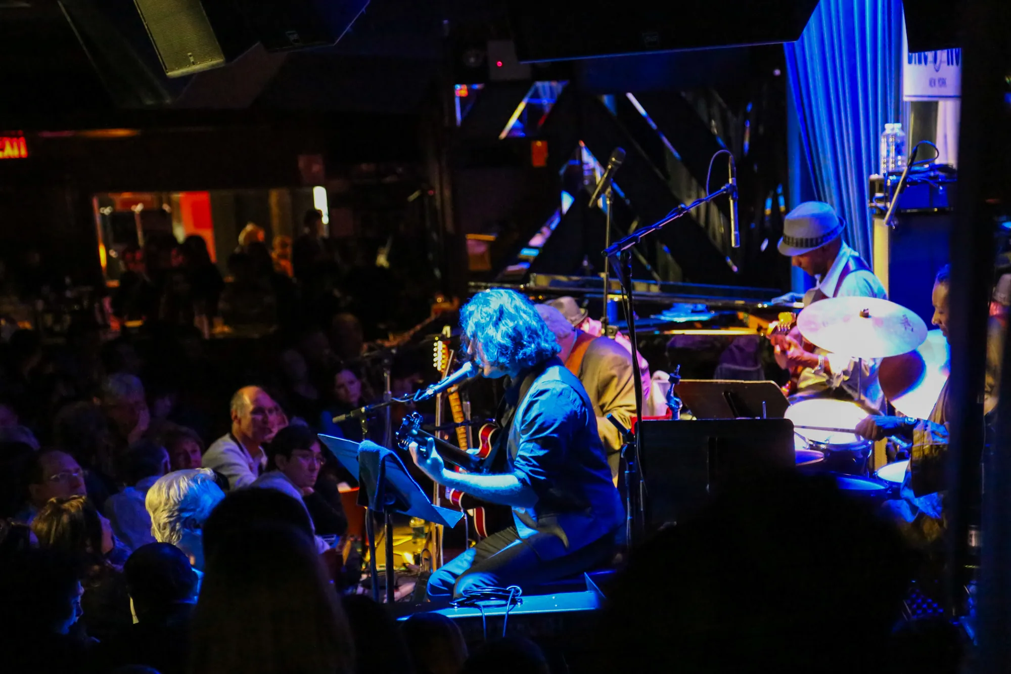 写真はアメリカ・ニューヨークのライブハウスで撮影されたものです。ステージ上ではバンドが演奏しており、ギター、キーボード、ドラムが確認できます。特にギターを弾きながら歌っている男性が中心に写っており、青い照明が当たっています。バンドの後ろには青いカーテンがあり、ステージを華やかに演出しています。客席は人で埋め尽くされており、観客は演奏に聴き入っている様子です。会場全体は暗く、ステージの照明が際立っており、ライブハウス独特の熱気と興奮が伝わってきます。