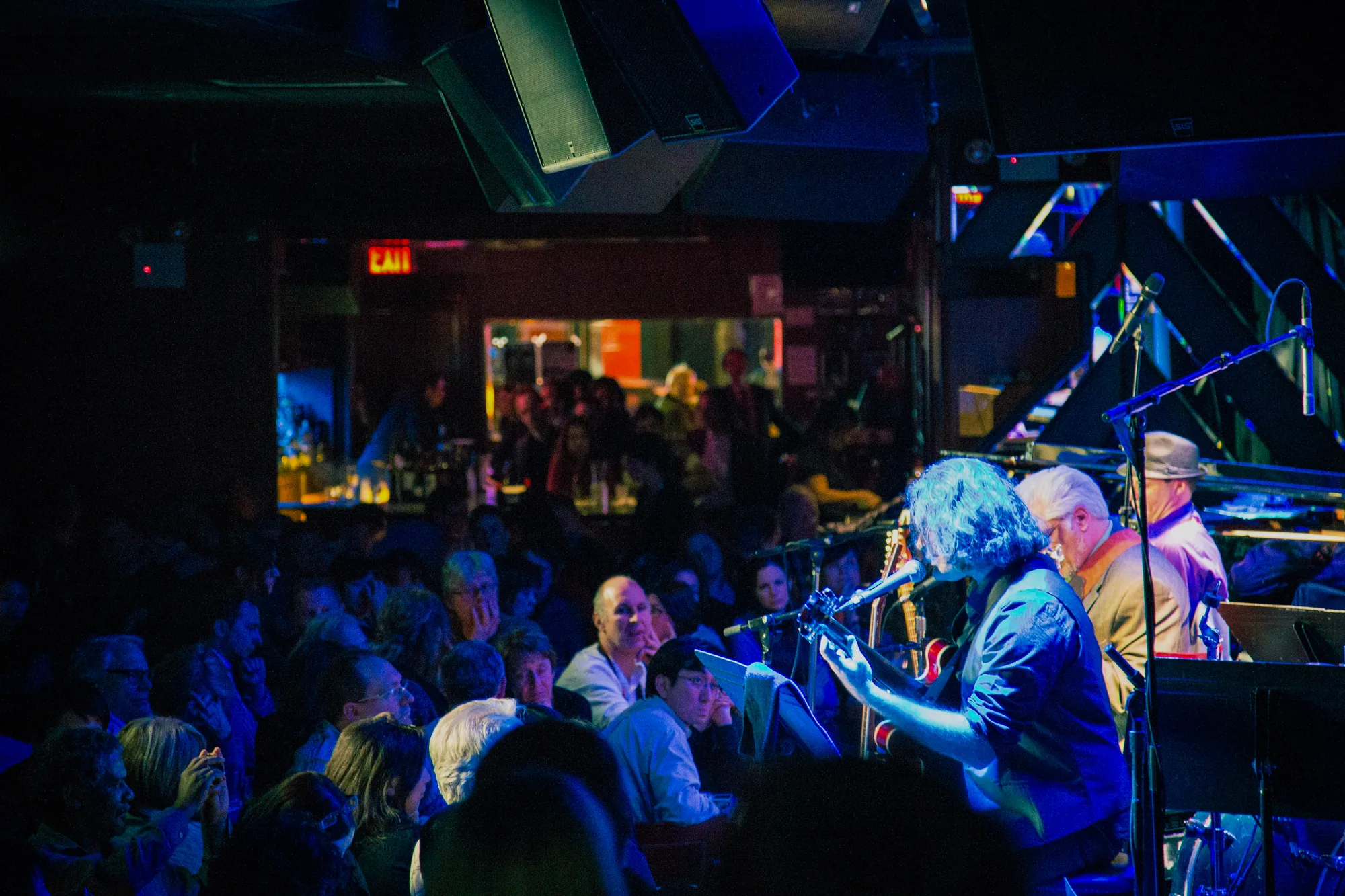 写真はアメリカ、ニューヨークのライブハウスで撮影されたものです。ステージ上では、ギターを弾きながら歌うミュージシャンと、ピアノを演奏するミュージシャンが演奏しています。ステージは青い照明で照らされており、ギターを弾くミュージシャンは青い光を浴びています。観客席はほぼ満席で、多くの人が演奏に耳を傾けています。観客の年齢層は高めで、落ち着いた雰囲気です。会場全体は薄暗く、天井には大型スピーカーが設置されています。奥にはバーカウンターが見え、EXITサインが光っています。全体的に、ニューヨークの都会的な雰囲気と、大人のための洗練されたライブエンターテイメントの雰囲気が感じられます。