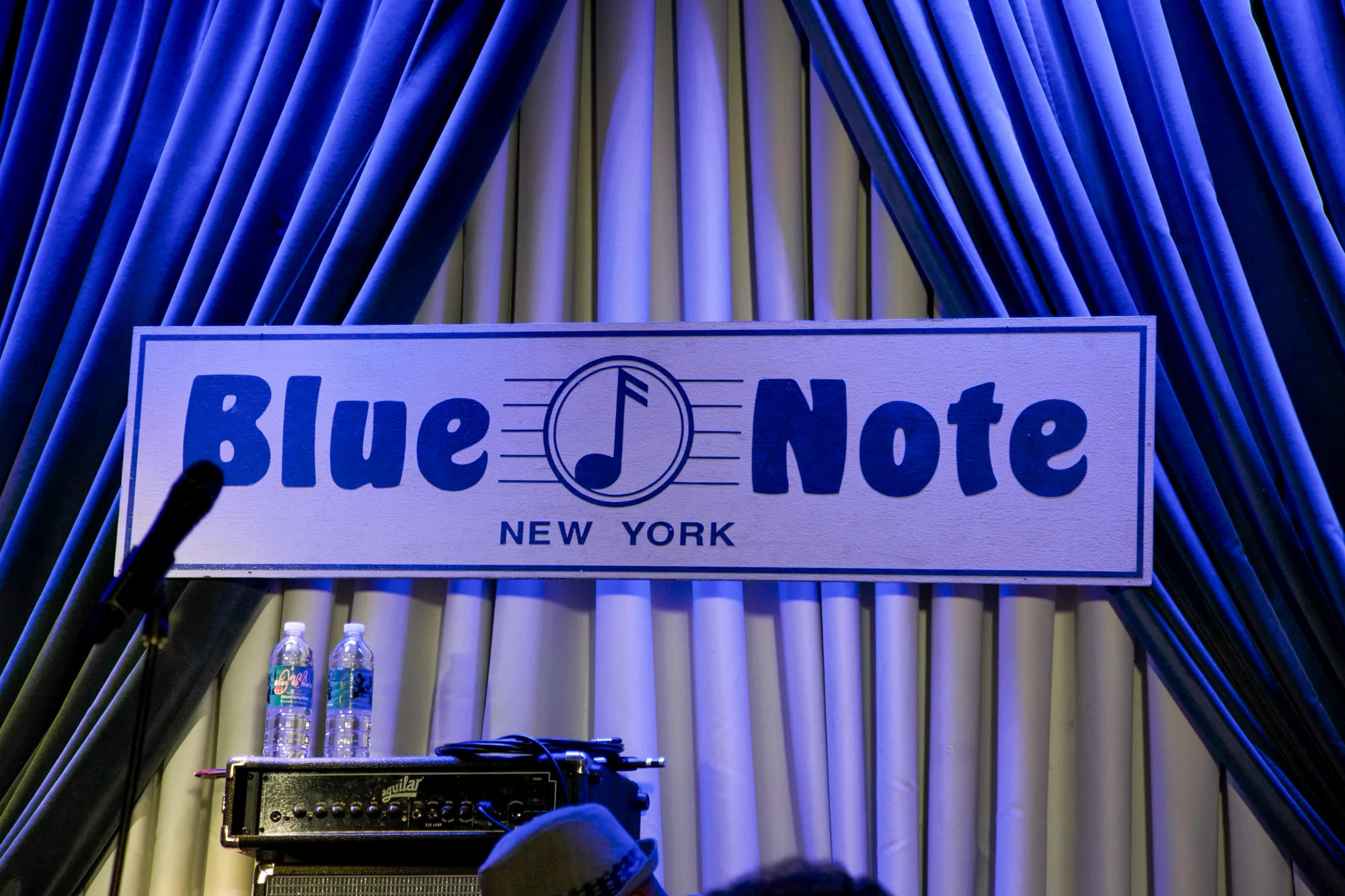 この写真は、アメリカ合衆国ニューヨークにある有名なジャズクラブ、ブルーノートの舞台を捉えたものです。中央に「Blue Note NEW YORK」と書かれた看板が掲げられており、そのロゴである音符のマークが目を引きます。看板の背後には、薄いグレーと濃いブルーのドレープが豪華に飾られ、舞台に独特の雰囲気を与えています。看板の下には、アンプやケーブル、ミネラルウォーターのボトルが置かれており、演奏の準備が整っている様子が伺えます。写真には、観客の一部も写っており、これから始まるであろう演奏への期待感が伝わってきます。全体的にブルーの照明が使用されており、落ち着いた大人の空間を演出しています。