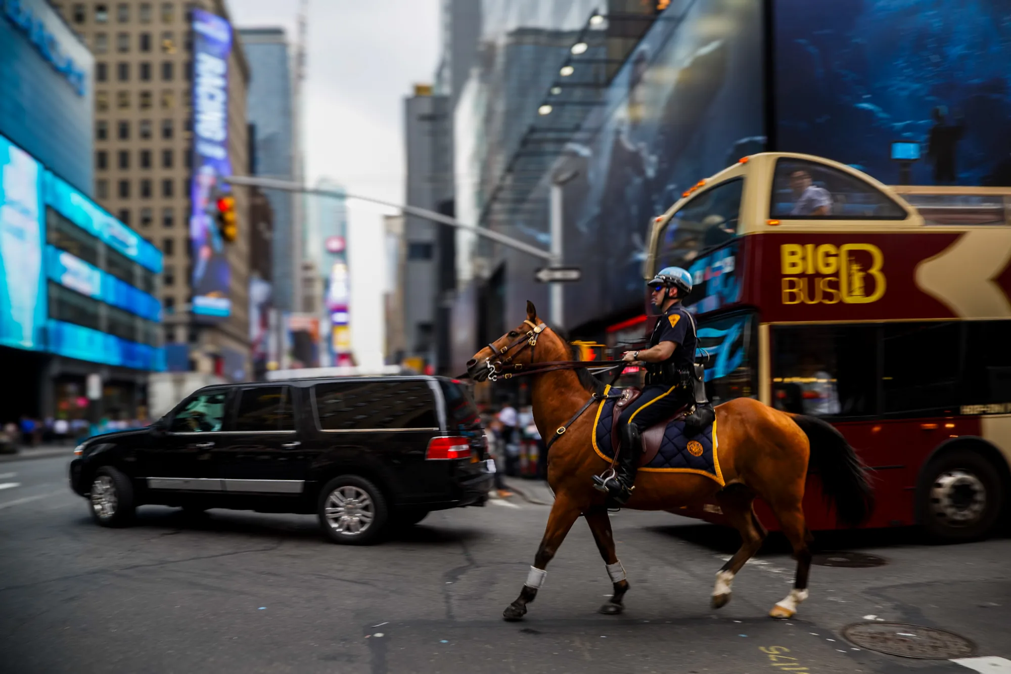 写真はアメリカ合衆国、ニューヨークの賑やかな通りで撮影されたものです。中央には、栗毛の馬に跨った騎馬警官が写っています。警官はヘルメットを被り、警察の制服を着ています。馬は歩いており、その足元には舗装された道路が見えます。背景には高層ビルが立ち並び、巨大なデジタル広告が輝いています。赤い二階建て観光バス「BIG BUS」が馬の横を通り過ぎようとしています。左側には黒いSUV車が写っており、全体的に都市の喧騒が感じられる雰囲気です。写真全体にモーションブラーがかかっており、躍動感が出ています。