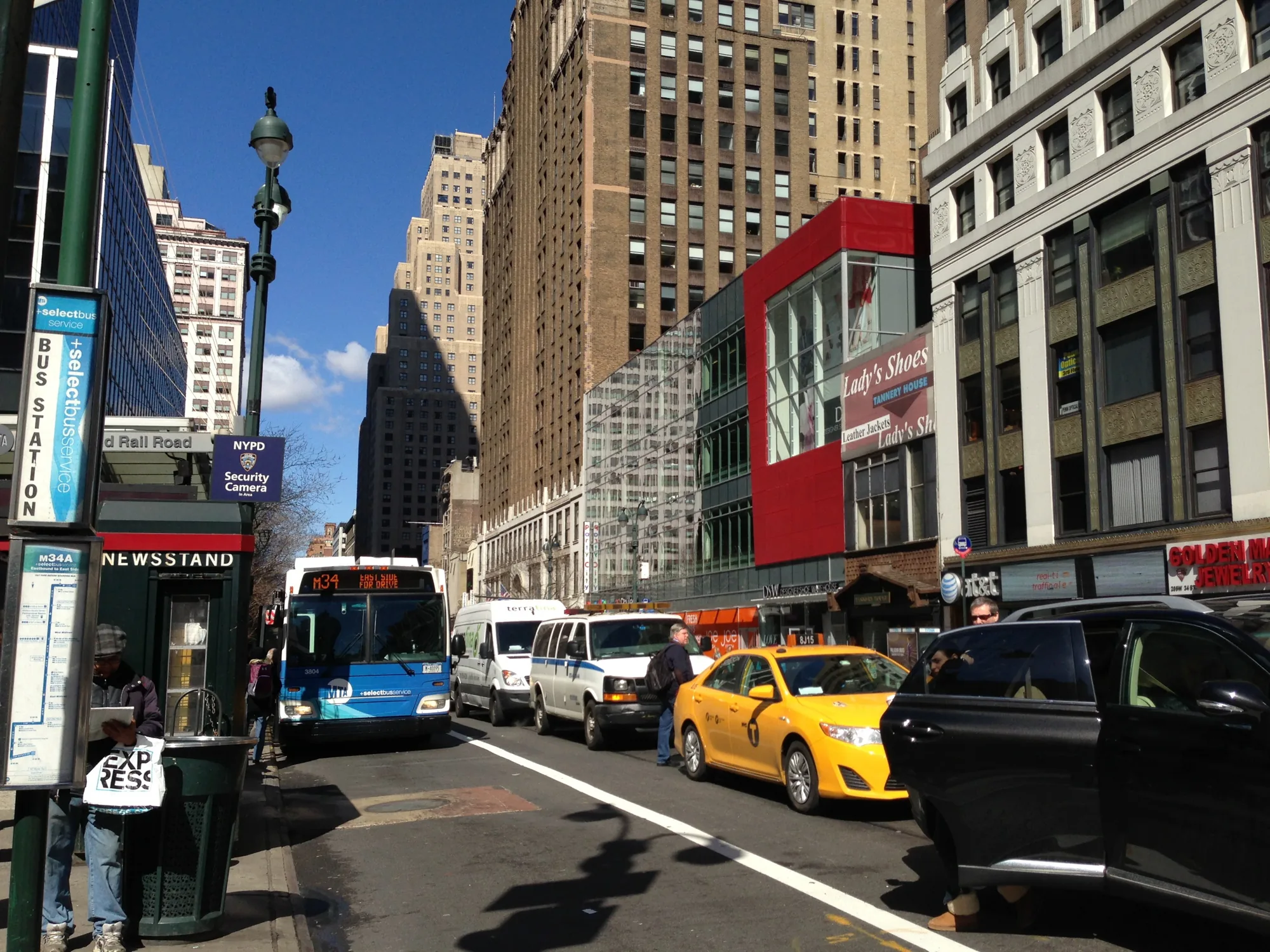 ニューヨークの街中で撮影された写真です。晴れた日の昼間で、青い空が広がっています。道路にはバス、タクシー、バンなどの車両が並んでいます。特に目立つのは、鮮やかな黄色のタクシーです。歩道にはバス停があり、ニューススタンドの横で男性が何かを読んでいます。背景には高層ビルが立ち並び、都会的な風景を形成しています。建物の壁には、店舗の看板や広告が見られます。全体的に活気があり、ニューヨークの日常的な風景を捉えた写真です。