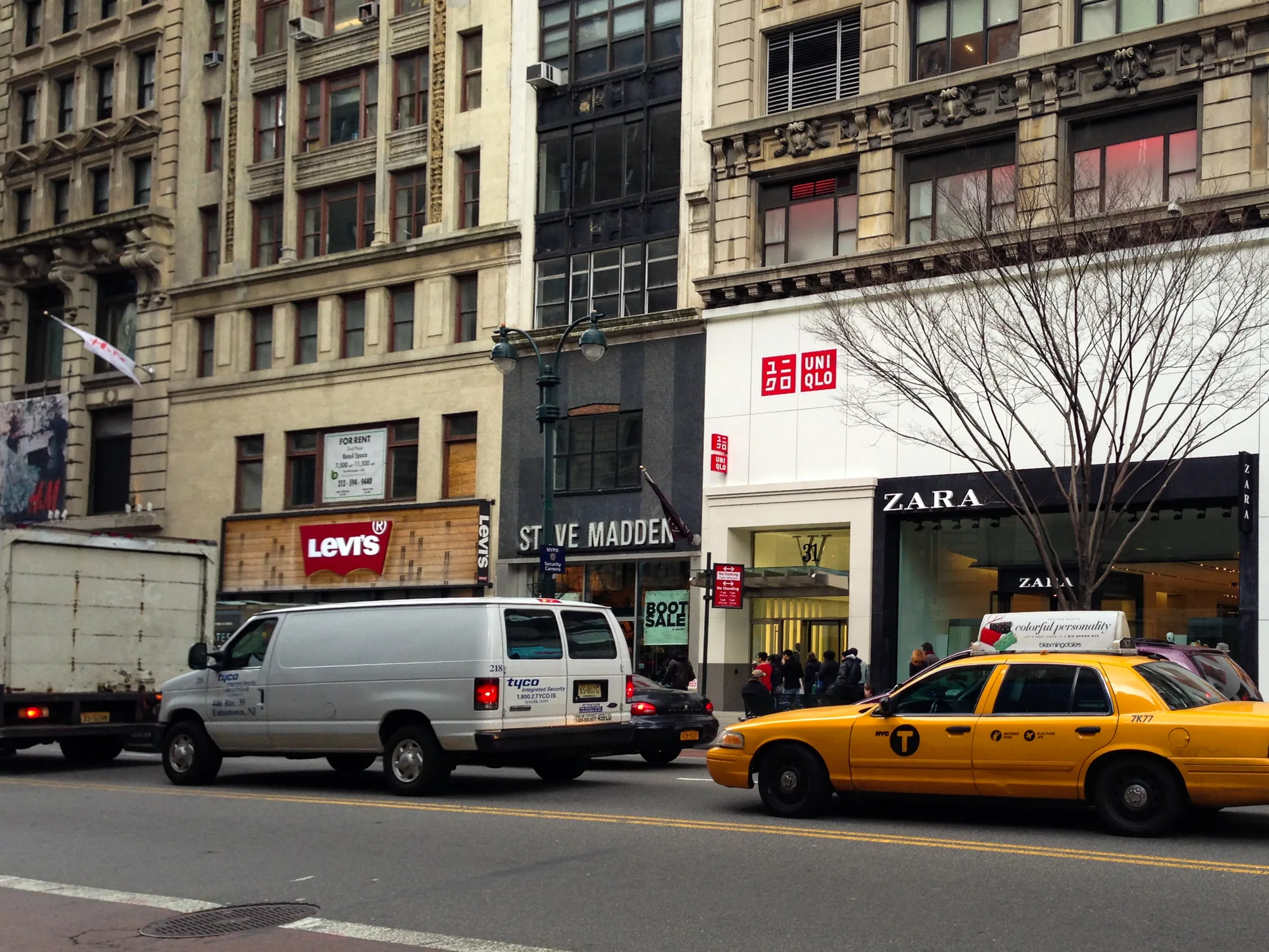 写真はアメリカ合衆国ニューヨーク市の街並みを捉えたものです。画面中央には道路が走り、その上を白いバンと、ニューヨーク名物の黄色いタクシーが走行しています。タクシーの屋根には広告が載っています。道路の反対側には複数の店舗が並んでおり、Levi's、Steve Madden、UNIQLO、ZARAなどのブランド名が確認できます。店には買い物客が出入りしており賑わいが見られます。背景には歴史を感じさせる石造りの建物がそびえ立ち、ニューヨークの都会的な雰囲気を醸し出しています。画面全体はやや曇り空で、落ち着いた色調となっています。