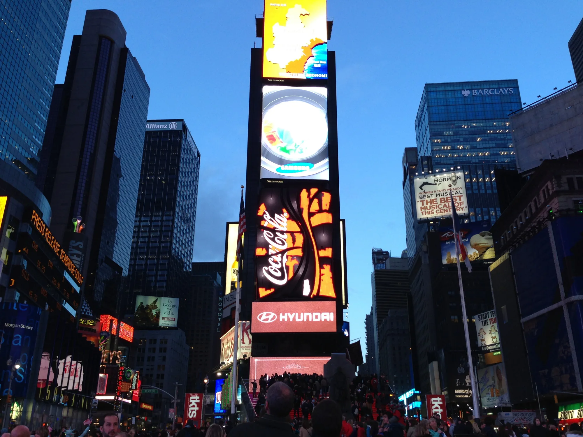 この写真はアメリカ合衆国ニューヨークのタイムズスクエアで撮影されたものです。夕暮れ時の空を背景に、高層ビル群がそびえ立ち、その間に巨大なデジタル広告がひしめき合っています。一番目立つ広告は、コカ・コーラと現代自動車（HYUNDAI）のものです。また、サムスンの広告やミュージカル「モルモン書」の広告も見られます。  広場は多くの人々で賑わっており、観光客や地元の人々が入り混じって歩いています。広告の光が人々の顔を照らし、活気に満ちた雰囲気を醸し出しています。写真全体は、タイムズスクエアのエネルギーと興奮を捉えており、ニューヨークの象徴的な風景を伝えています。