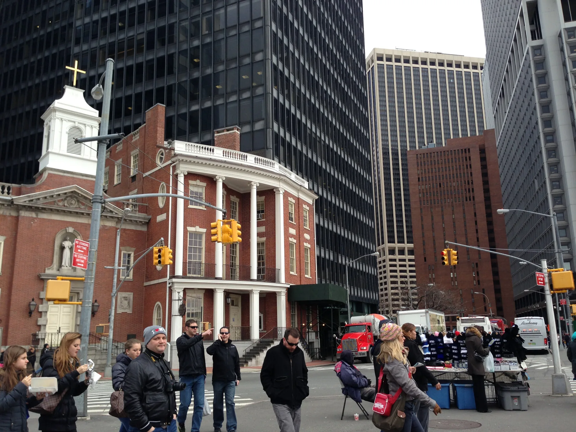 この写真はアメリカ、ニューヨークで撮影された街並みの風景です。画面左側にはレンガ造りの歴史を感じさせる建物があり、上部には十字架が掲げられています。看板には「NO STANDING ANYTIME」と書かれています。その背景には、ガラス張りの高層ビルがそびえ立ち、新旧の建築様式が混在している様子が伺えます。道路には歩行者が何人かいて、横断歩道を歩いています。露店も出ており、帽子やマフラーなどを販売しているようです。全体的に曇り空で、落ち着いた雰囲気の写真です。