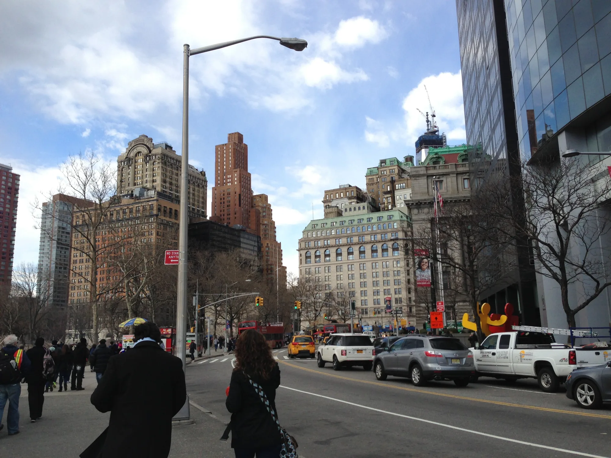 写真は、アメリカ合衆国ニューヨークで撮影された、都会の街並みを捉えたものです。歩道には多くの人々が歩いており、道路には様々な種類の車が走っています。背景には、レンガ造りや石造りの高層ビルが立ち並び、歴史を感じさせる重厚な雰囲気を醸し出しています。建設中の建物も見られ、都市が常に変化し続けている様子が伺えます。空には白い雲が浮かび、青空が広がっています。全体的に、活気があり、洗練された都会の雰囲気が感じられます。