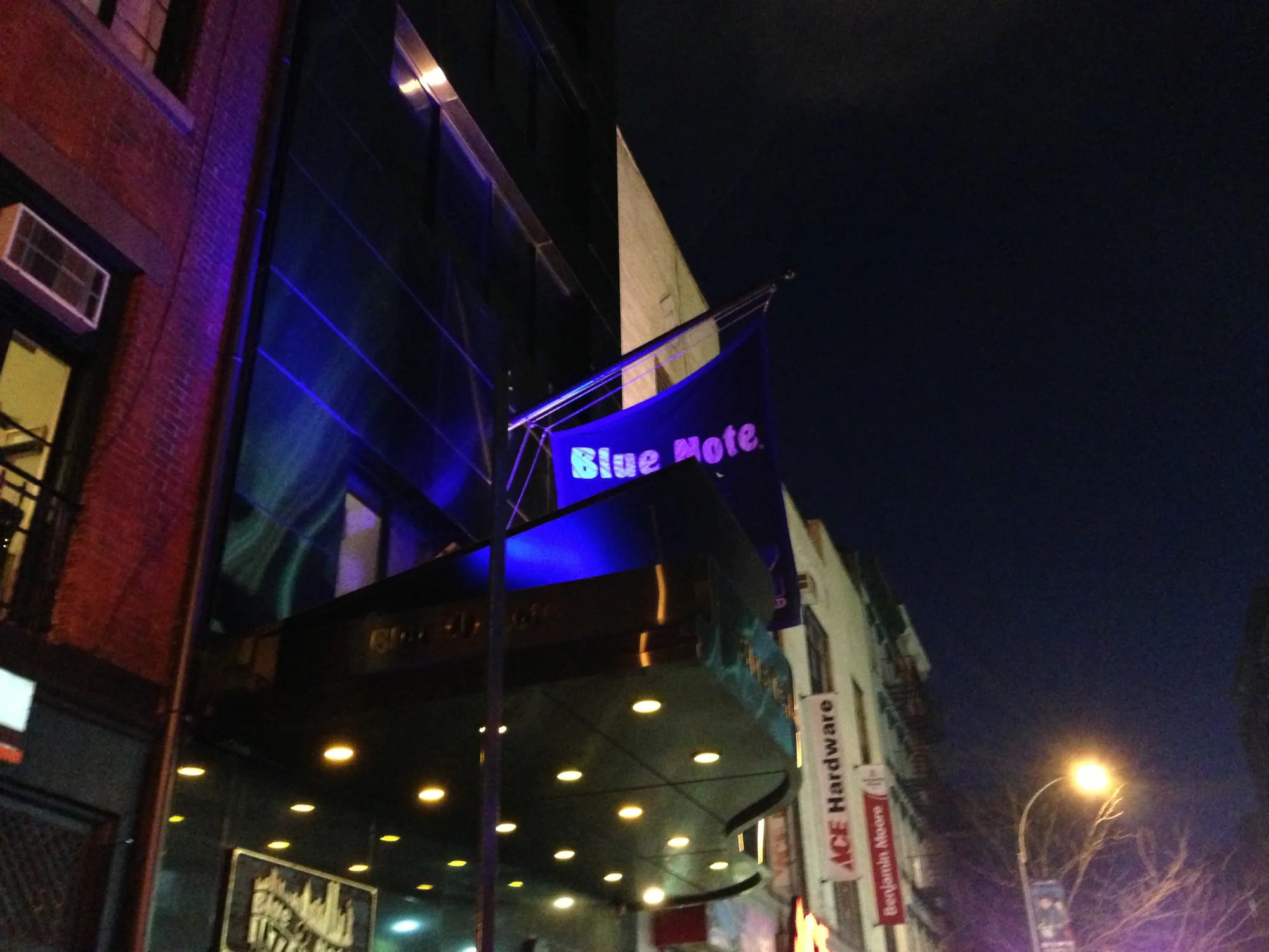 夜のニューヨークで撮影された写真です。Blue Noteの建物が大きく写っており、青い照明が印象的です。建物の入り口にはAce Hardwareの看板も見えます。街灯が夜道を照らし、活気のある雰囲気を醸し出しています。