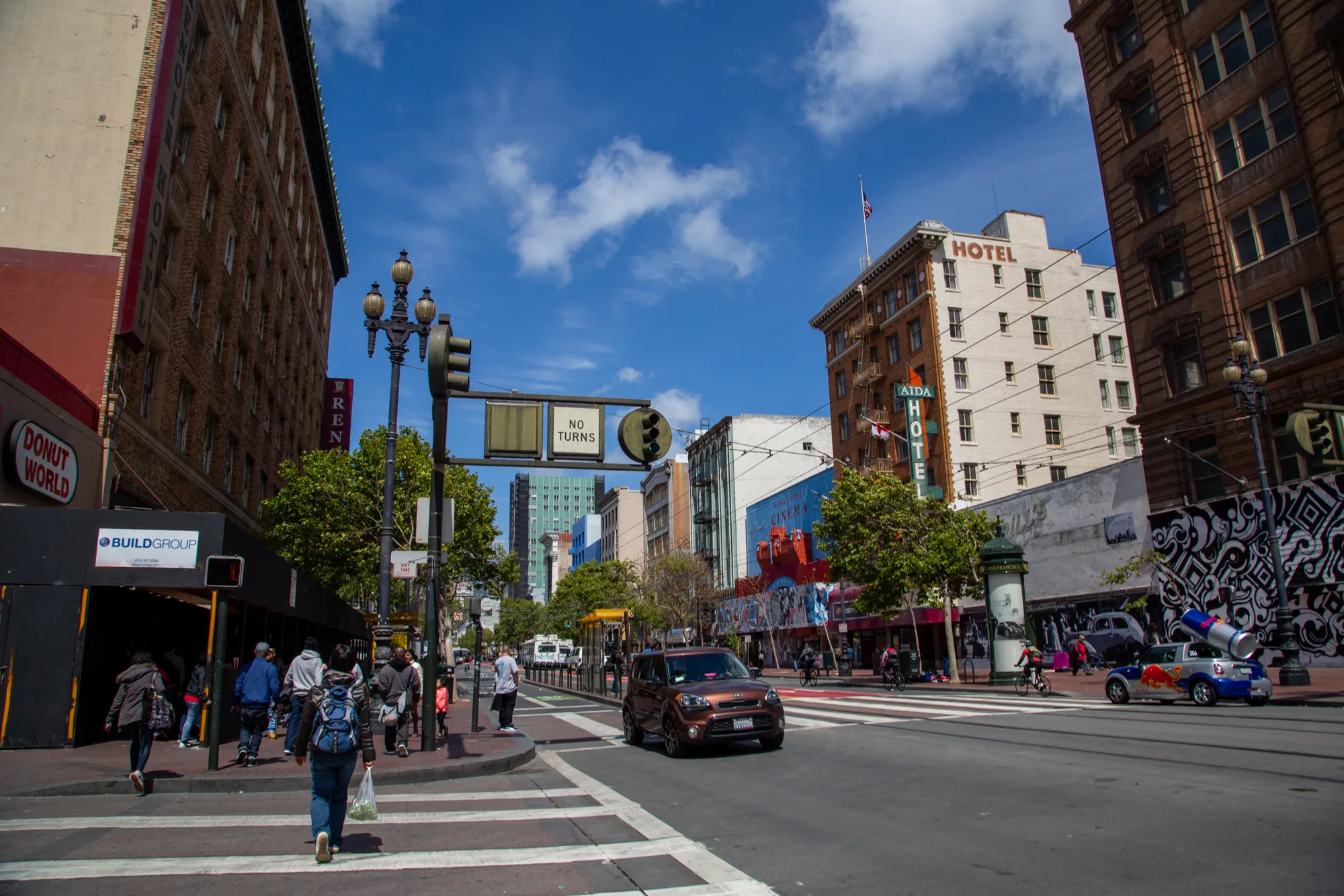 写真はアメリカ合衆国サンフランシスコで撮影された街の風景です。青空が広がり、白い雲が浮かんでいます。画面中央には、茶色の車が走行しており、手前の横断歩道には歩行者が数名います。道路の左側には「DONUT WORLD」という看板のある店舗があり、その隣には工事現場のような場所があります。道路の右側には「AIDA HOTEL」と書かれた建物が見えます。背景には高層ビル群が立ち並び、都会的な景観が広がっています。写真全体を通して、活気のある街の雰囲気が感じられます。赤ブルの宣伝カーも写っています。