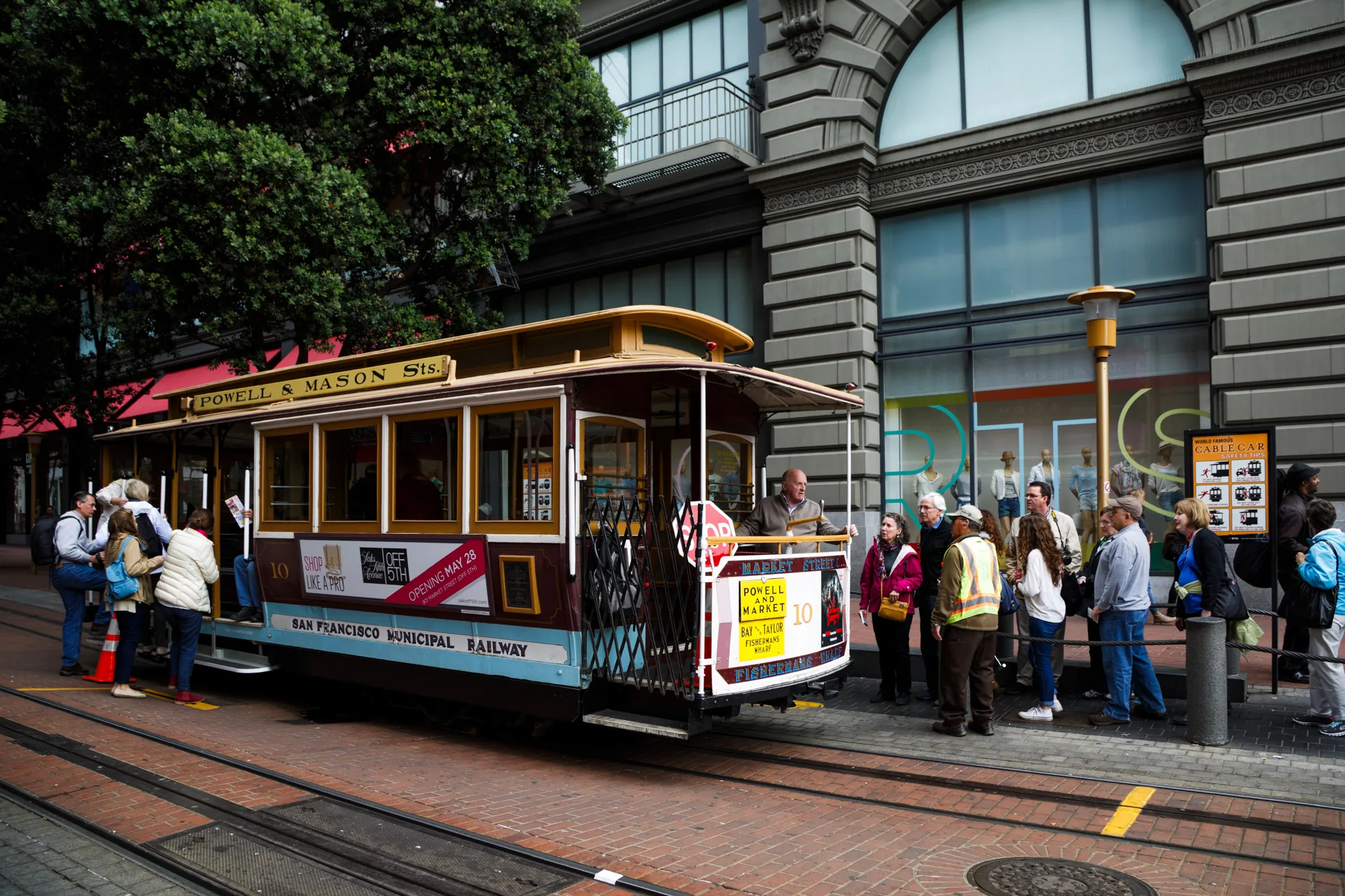写真はアメリカ合衆国サンフランシスコで撮影されたもので、街中を走るケーブルカーが写っています。ケーブルカーは Powell & Mason St.の路線で、外装は茶色と水色で塗装されています。「SAN FRANCISCO MUNICIPAL RAILWAY」と書かれています。ケーブルカーには多くの人が乗り込んでおり、またケーブルカーの前や横にも人が集まっています。人々はカジュアルな服装をしており、観光客が多いようです。背景にはレンガ敷きの道路、線路、そして重厚な石造りの建物が写っています。建物の窓にはマネキンが飾られており、お店であることがわかります。全体的にやや曇り空で、落ち着いた雰囲気の写真です。