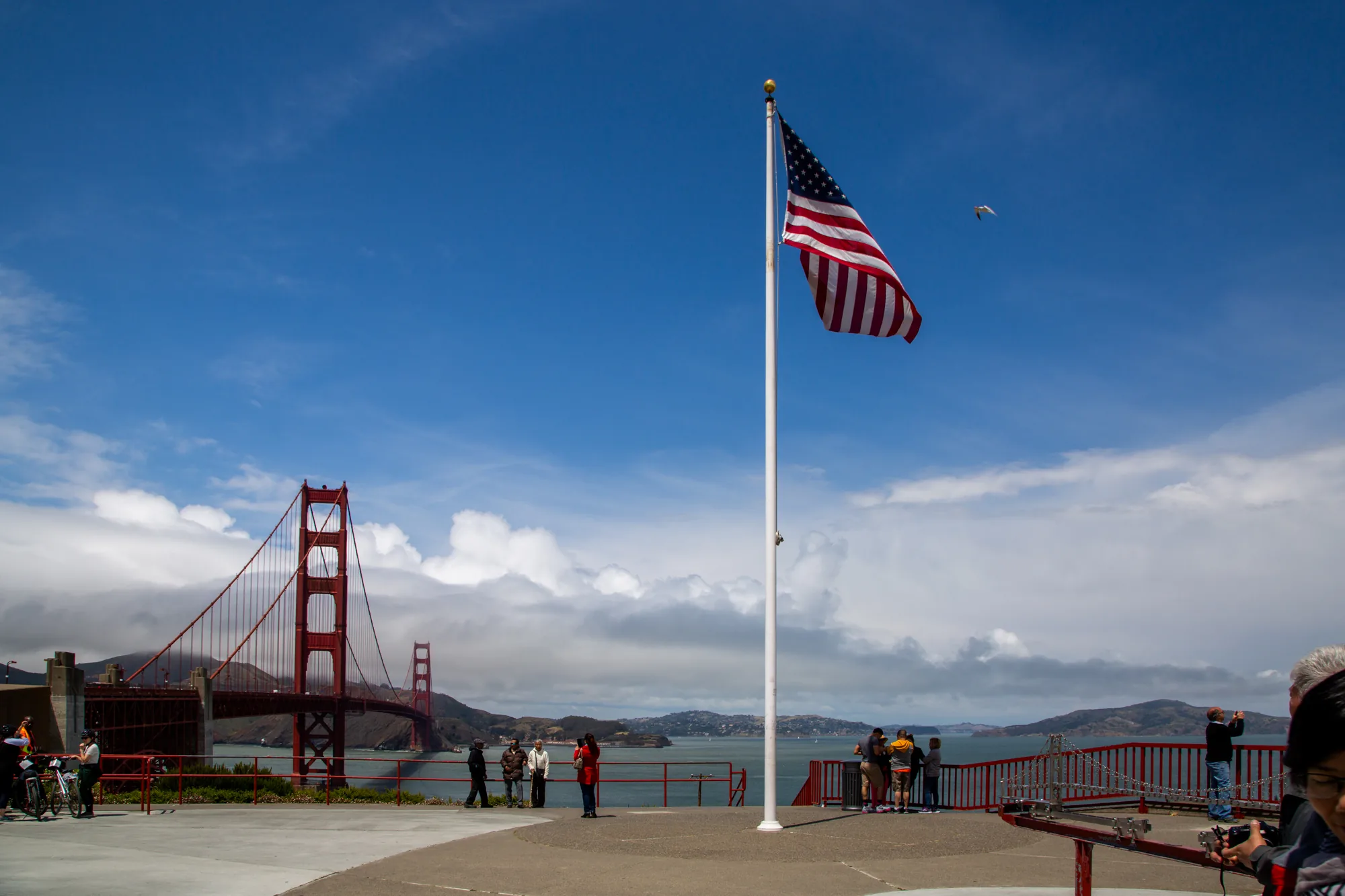 写真には、アメリカのサンフランシスコにある金門橋が写っています。手前にはアメリカの国旗が掲げられており、空には白い雲が浮かんでいます。橋のたもとには、観光客らしき人々が景色を眺めたり、写真を撮ったりしています。画面右端には、カメラを構える人物の頭部が見切れて写っています。明るい日差しの中で、アメリカらしい風景が広がっています。