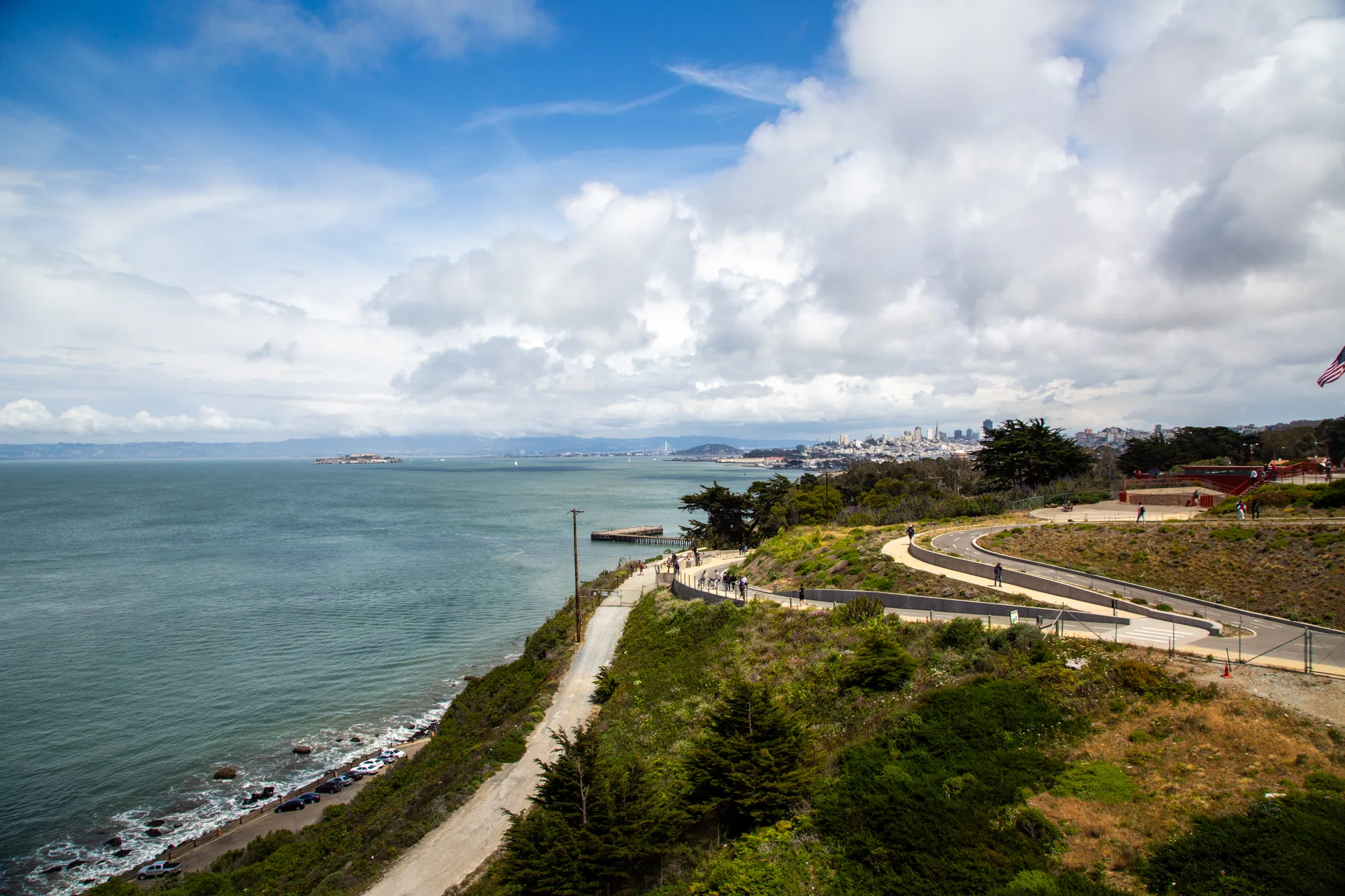 写真は、アメリカ合衆国のサンフランシスコで撮影された風景を捉えています。手前には、緑豊かな丘陵地帯が広がり、様々な種類の木々や草が生い茂っています。丘の斜面には、人が歩けるように整備された道があり、何人かの人々が散策を楽しんでいます。  丘の下には海岸線が広がり、荒々しい波が岸に打ち寄せています。海岸沿いには、車が数台駐車しているのが見えます。海面は穏やかで、遠くにはアルカトラズ島が浮かんでいます。さらに遠方には、サンフランシスコの市街地が、霧にかすんで見えています。  空には、白い雲が大きく広がり、青空とのコントラストが美しいです。全体的に、自然の雄大さと都市の景観が調和した、爽やかな印象の写真です。右端にはアメリカ国旗が一部写っています。
