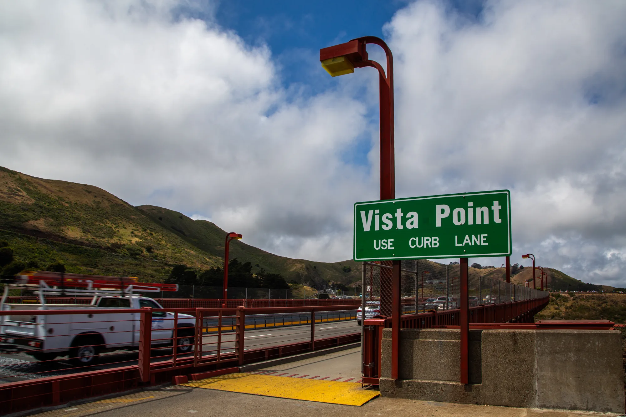 サンフランシスコで撮影されたと思われる写真です。手前には緑色の「Vista Point USE CURB LANE」と書かれた道路標識が立っています。標識は赤いポールに取り付けられており、上部には黄色い照明が設置されています。  写真の奥には橋が伸びており、赤い柵が特徴的です。橋の上を車が走行しており、左方向へ白いトラックが走っているのが見えます。橋の向こうには草木に覆われた丘陵地帯が広がっており、空には白い雲が浮かんでいます。全体的に明るい雰囲気で、晴れた日の様子が伝わってきます。橋の構造物や標識のデザインから、アメリカの風景であることが伺えます。