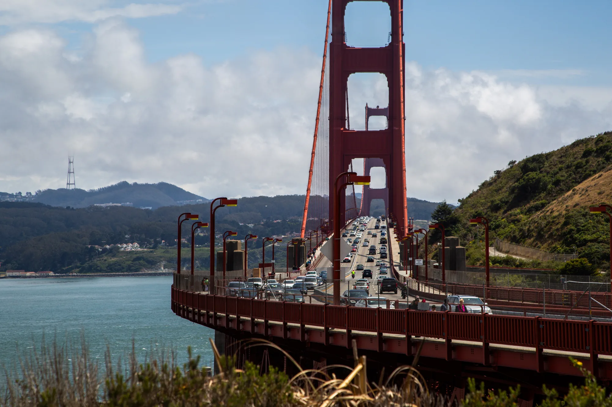 この写真は、アメリカのサンフランシスコにある金門橋を捉えたものです。カメラは橋の一部をフレームに収め、橋の上を走る車の流れを見ることができます。赤い橋の構造物が前景にあり、その上に交通量の多い道路が広がっています。橋の向こうには、青い海、遠くの山々、そして白い雲が浮かぶ空が広がっています。山の上には Sutro Tower が見えます。橋の歩道には人が歩いているのが見えます。全体的に、活気がありながらも落ち着いた雰囲気の写真です。