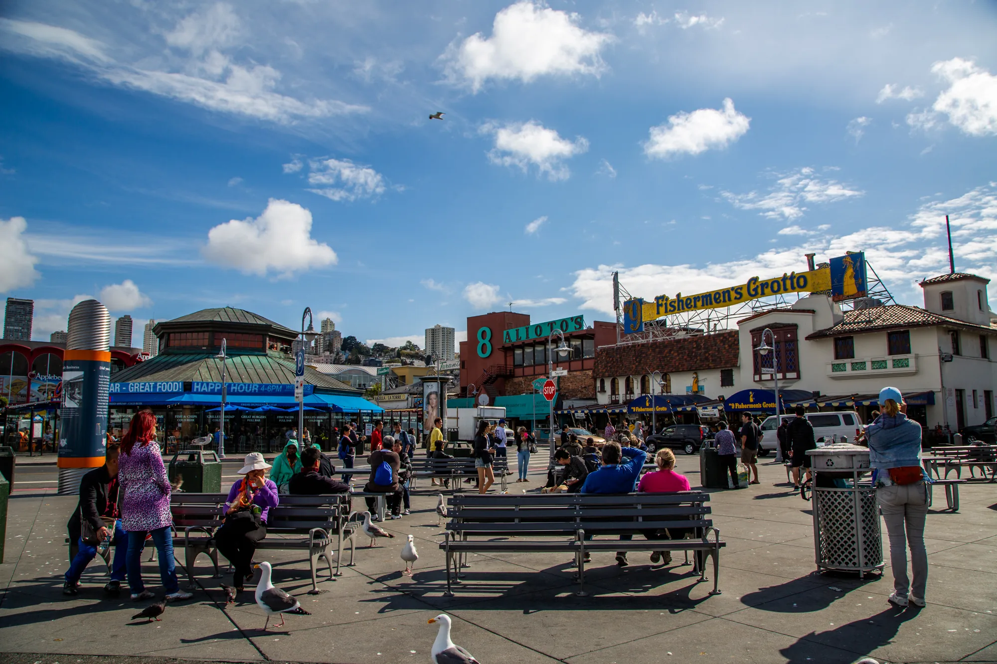 写真はアメリカ、サンフランシスコで撮影されたものです。場所は有名な観光地であるフィッシャーマンズワーフ周辺だと思われます。青い空には白い雲が浮かび、日差しが明るい日中の様子が写されています。  写真には、多くの人々が行き交う様子が写っており、観光客や地元の人々で賑わっていることがわかります。ベンチに座って休憩している人、歩いている人、景色を眺めている人など、様々な姿が見られます。  背景には、有名なレストラン「Alioto's」や「Fisherman's Grotto」の看板が見えます。これらの建物はサンフランシスコの象徴的な存在であり、観光客に人気があります。また、遠景にはサンフランシスコの街並みも見え、高層ビルが立ち並んでいます。  前景には、地面を歩き回るカモメが写っており、港町らしい雰囲気を醸し出しています。全体的に、活気があり、楽しげな観光地の様子が伝わってくる写真です。