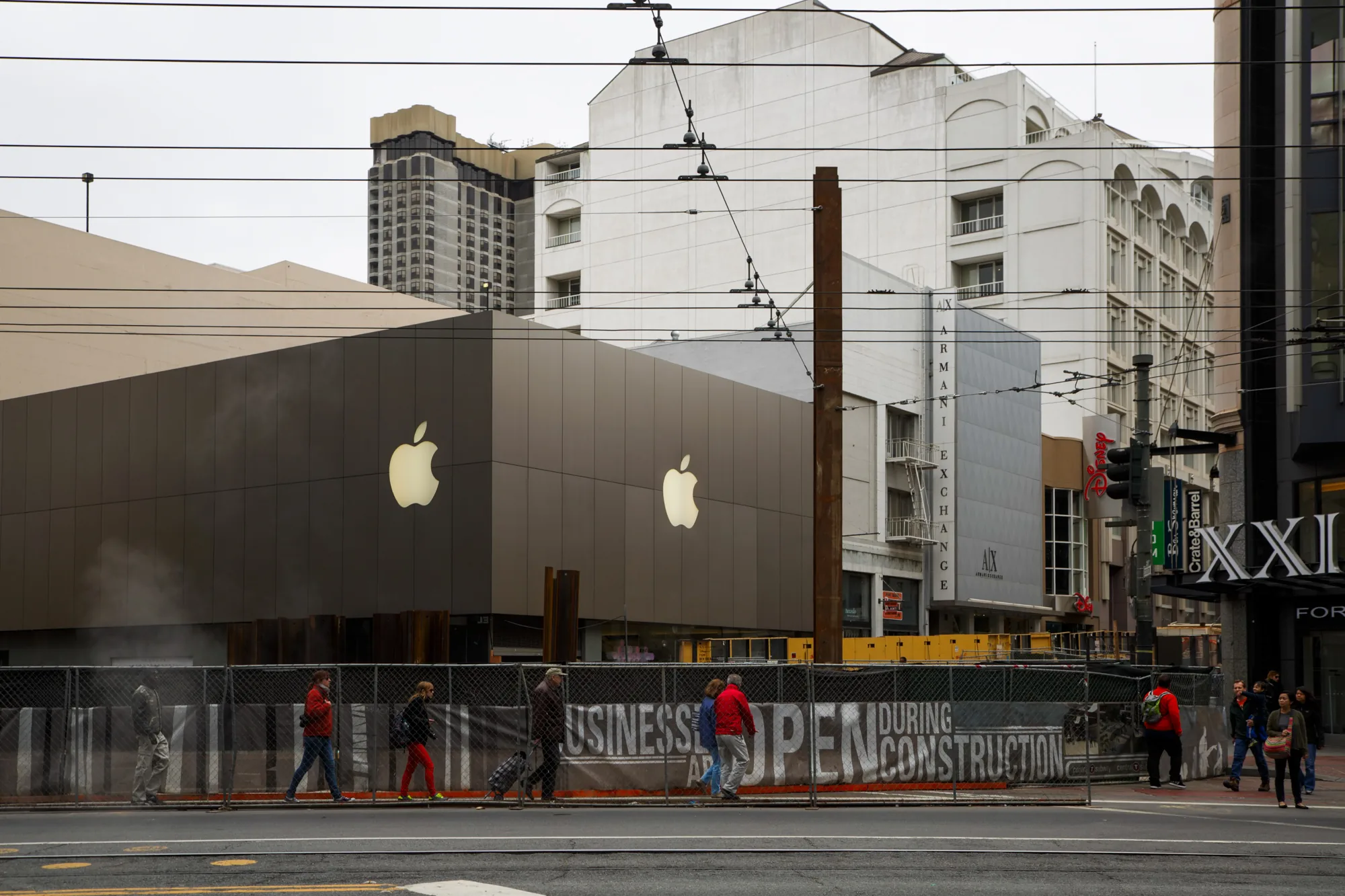 サンフランシスコの街中で撮影された写真です。手前には歩道を歩く人々が写っており、その奥には工事中のアップルストアが見えます。建物の外観はダークグレーで、大きく光るアップルのロゴが印象的です。工事現場はフェンスで囲われており、「BUSINESSES ARE OPEN DURING CONSTRUCTION（営業中）」というメッセージが書かれています。写真には、アップルストアの他に、ARMANI EXCHANGEやCrate & Barrelなど、複数のブランドの看板が見られます。電線が空を横切り、周囲には高層ビルや古い建築様式の建物が混在しています。全体的に曇り空で、ややくすんだ色調が、都市の落ち着いた雰囲気を醸し出しています。