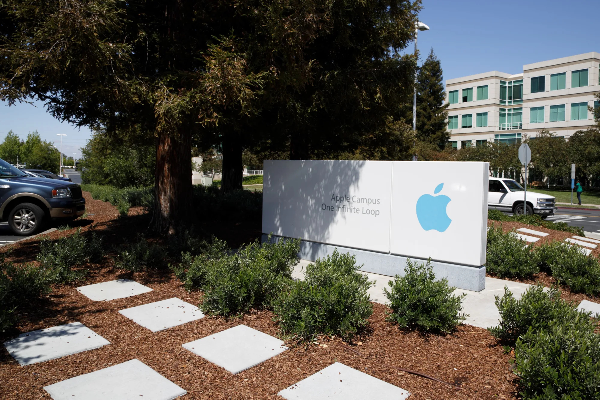 アメリカ合衆国カリフォルニア州サンフランシスコで撮影された写真です。写真はApple Campusの入口付近を捉えています。画面中央には、白地の大きな看板があり、Appleのロゴと「Apple Campus One Infinite Loop」という文字が薄いグレーで印字されています。看板の周りには、木々や低木が植えられており、地面はウッドチップで覆われています。白い四角い石が飛び石のように配置されています。  写真の左側には、青い車の一部が見えます。画面奥には、オフィスビルとその他の車両が写っています。空は晴れており、全体的に穏やかな雰囲気です。看板には木々の影が落ちています。