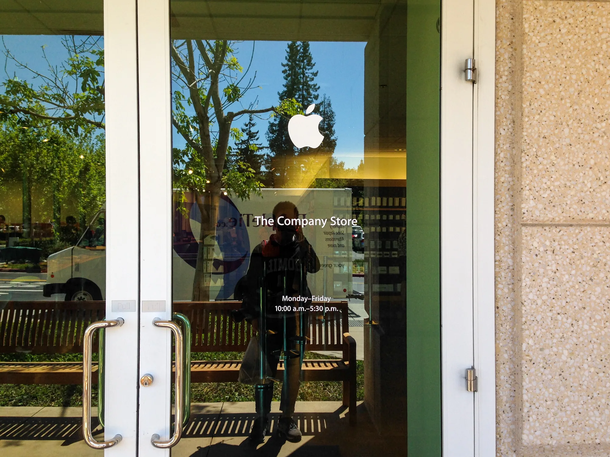 写真は、アメリカのサンフランシスコにあるApple社のストアの入り口を撮影したものです。白いドアとガラス窓が特徴的で、窓にはAppleのロゴと「The Company Store」という文字がプリントされています。また、営業時間が「Monday-Friday 10:00 a.m.-5:30 p.m.」と記載されています。  ドアの向こうには、通りの景色が反射して映っており、木々や空、白いバン、ベンチなどが見えます。また、ドアのガラスに映り込んでいる人物像も見えます。その人物はカメラを持っており、写真を撮っているようです。ドアの取っ手はシルバーで、清潔感のある印象を与えます。建物の外壁は、ベージュ色の石造りで、小さな斑点模様が入っています。全体的に明るく、穏やかな雰囲気の写真です。