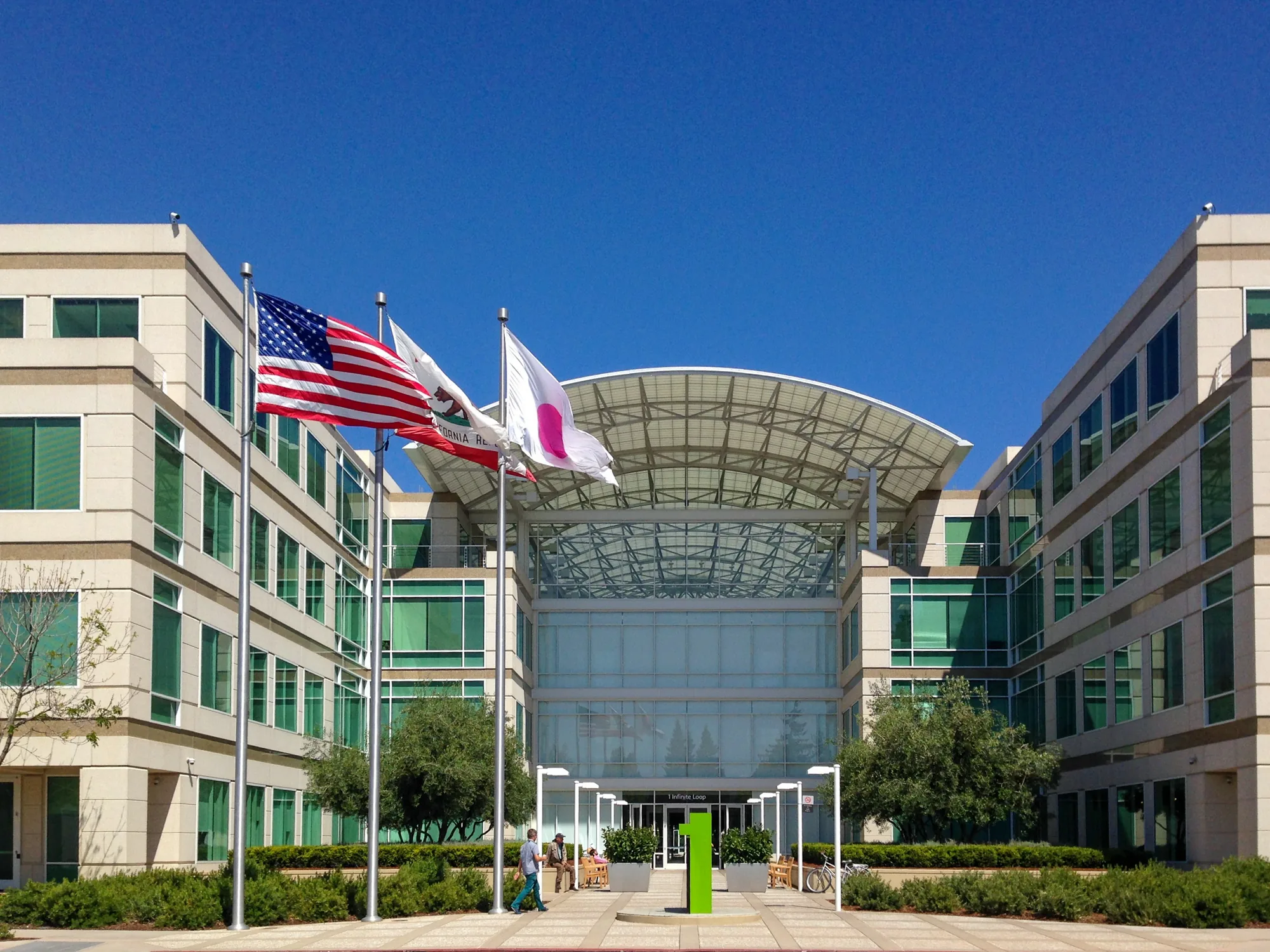 写真は、アメリカのサンフランシスコにあるオフィスビルを捉えたものです。快晴の青空の下、建物は左右対称に構成されており、モダンなデザインが特徴的です。中央にはガラス張りのエントランスがあり、屋根は半円状の金属製構造物で覆われています。エントランスの前には緑色のモニュメントが立っており、その奥にはベンチに座って休憩している人々が見えます。建物の前には、アメリカ国旗、カリフォルニア州旗、そして日の丸が掲げられています。周囲には緑豊かな植栽が施され、整然とした景観を作り出しています。全体的に、清潔感があり、洗練された雰囲気が漂っています。住所からおそらくAppleの旧本社であるInfinite Loopと思われます。