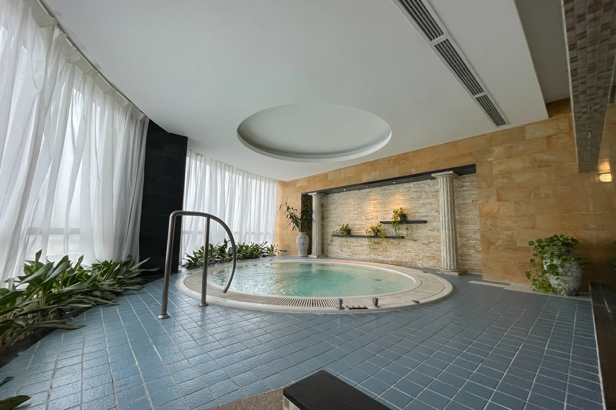 写真はベトナムのハノイで撮影されたスパの室内の様子を捉えています。中央には円形のジャグジーがあり、水面は穏やかに波打っています。ジャグジーの周囲は青いタイルで覆われており、清潔感があります。窓際には白いカーテンがかけられ、自然光が柔らかく差し込んでいます。壁の一部は石材で装飾され、柱の装飾や植物が配置されており、落ち着いた雰囲気を醸し出しています。天井には円形のデザインがあり、照明が組み込まれています。全体的に高級感があり、リラックスできる空間であることが伺えます。