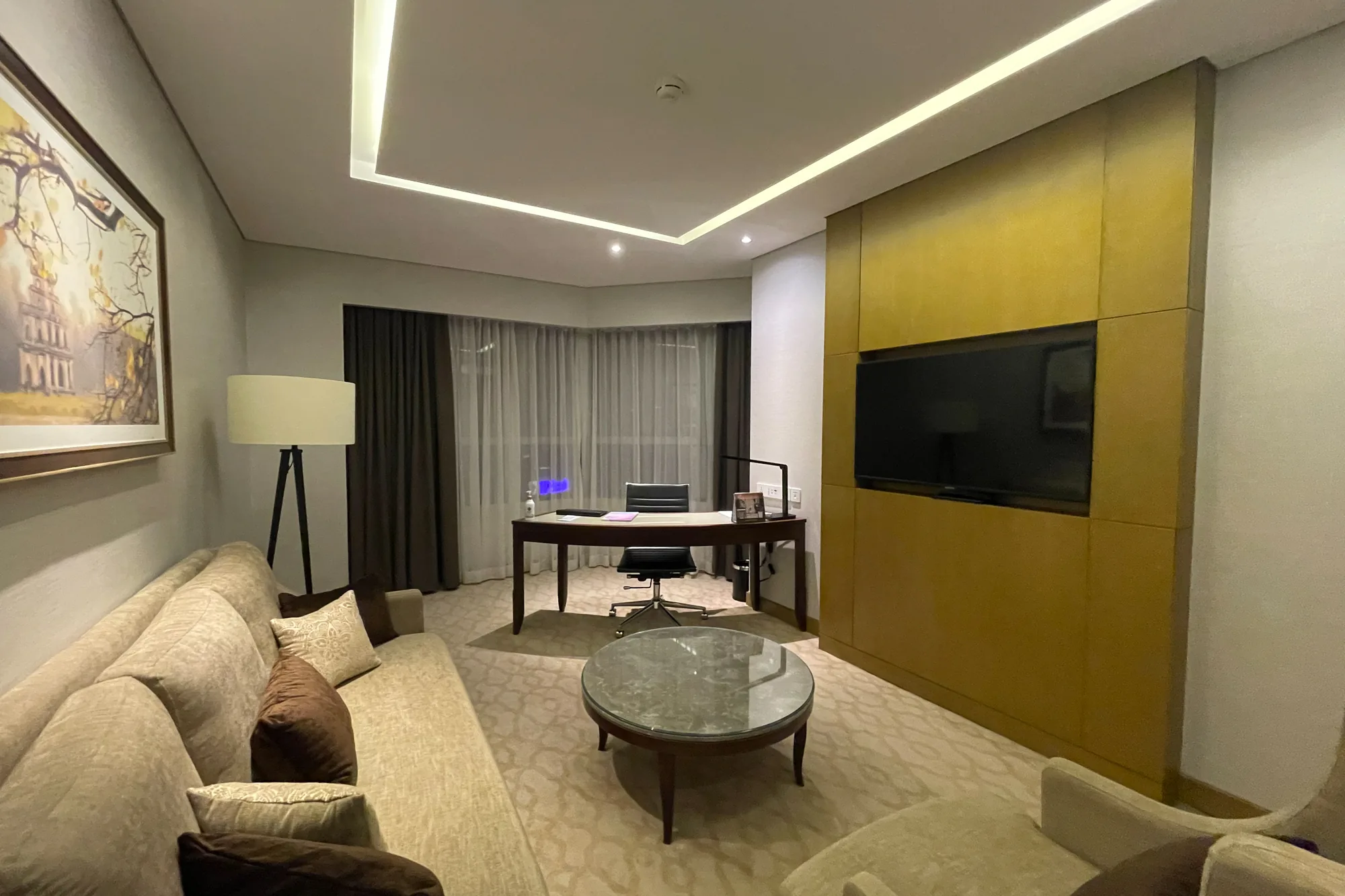 写真はベトナムのハノイで撮影された、ホテルの部屋の様子を捉えたものです。部屋はベージュとブラウンを基調とした落ち着いた色調でまとめられています。  画面左手には、ベージュ色のソファが置かれ、その上には茶色のクッションがいくつか配置されています。ソファのそばには、三脚のフロアランプが立っています。壁には額縁に入った絵画が飾られています。  部屋の中央には、丸いガラステーブルが置かれています。奥には、木製のデスクと椅子が設置され、デスク上にはデスクライトや小物が見えます。窓にはカーテンが引かれ、自然光が柔らかく差し込んでいます。  画面右手の壁には、大きなテレビが壁掛けされています。壁全体が木製のパネルで覆われており、照明が反射して光沢を帯びています。部屋全体は間接照明で照らされ、穏やかで快適な雰囲気を醸し出しています。ビジネスで利用するにも、リラックスするにも適した空間だと感じられます。