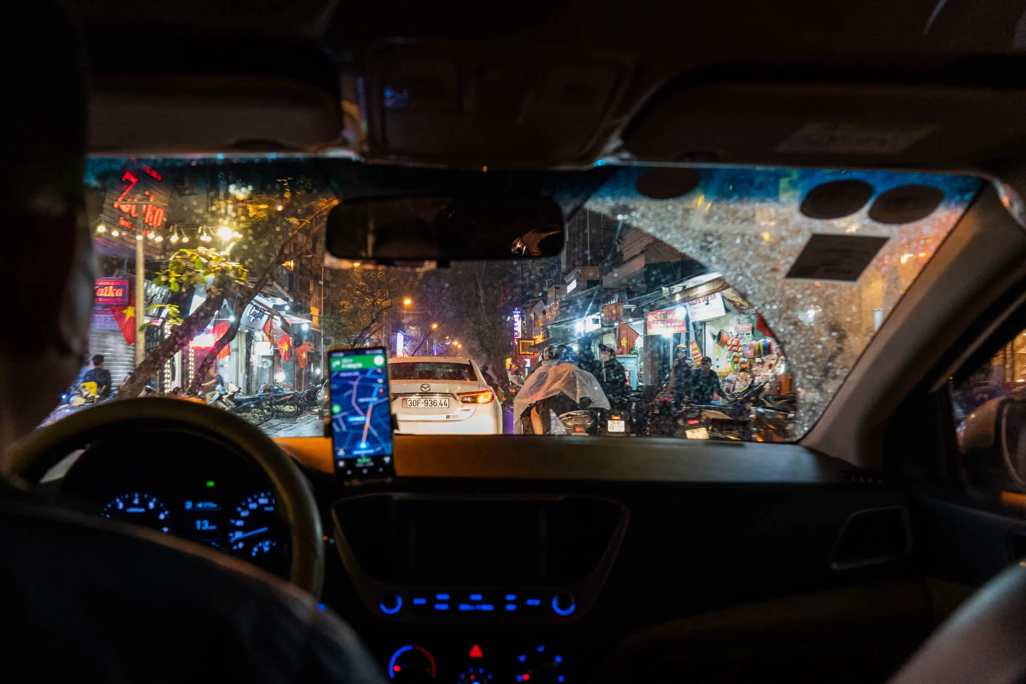 写真は、ベトナムのハノイで撮影された、車の運転席からの風景です。夜の街並みがフロントガラス越しに見え、雨が降っているため、ガラスには水滴が付着しています。ダッシュボードにはスマートフォンが設置されており、ナビゲーションアプリが表示されています。車のメーターパネルは青色に光っており、速度計や燃料計などが確認できます。フロントガラスの外には、バイクや人々が行き交う様子が写っており、通り沿いには小さなお店が並んでいます。ベトナムの国旗が掲げられているのが見えます。街灯の光やお店の明かりが夜の街を照らし、活気ある雰囲気を醸し出しています。