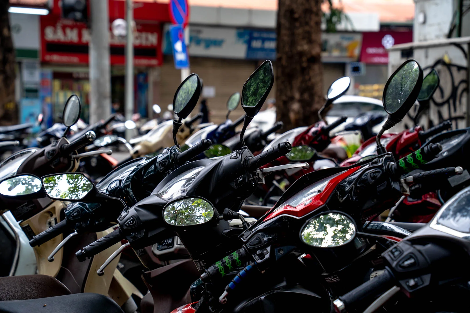 この写真はベトナムのハノイで撮影されたもので、大量のバイクが整然と駐車されている様子を捉えています。写真全体はバイクのハンドルやミラーで埋め尽くされており、ハノイの街におけるバイクの普及率の高さを物語っています。バイクの色や種類は様々で、赤、黒、ベージュ、青などが見られます。一部のバイクのミラーには周囲の木々や建物が反射しており、街の喧騒が感じられます。背景には店舗の看板や交通標識が写っており、ハノイの日常的な風景の一部を垣間見ることができます。全体的にやや曇りがちな天気であり、落ち着いた雰囲気の写真です。