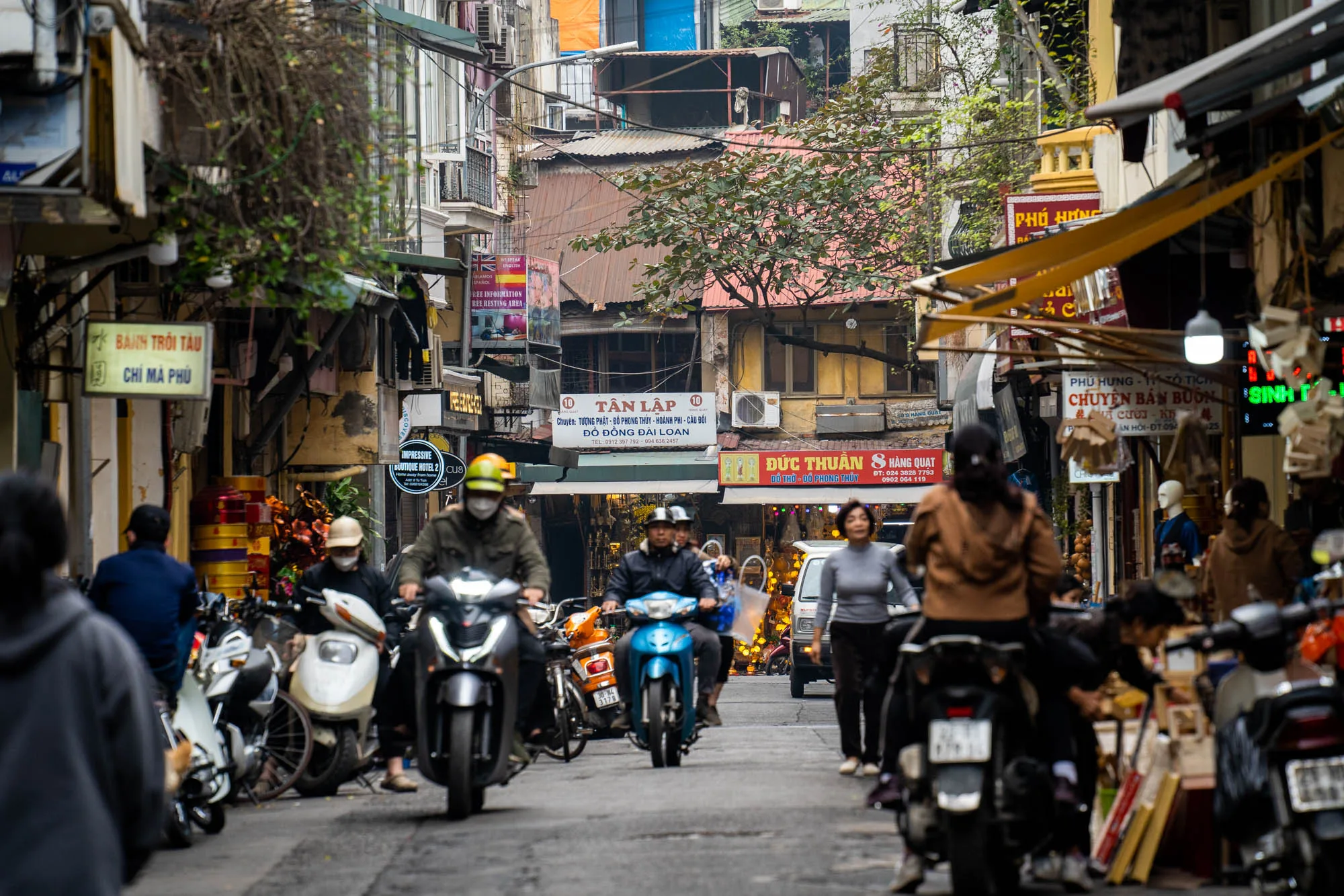 この写真はベトナムのハノイで撮影された、賑やかな路地の様子を捉えています。道の中心部には、ヘルメットを被った人々が運転する多数のバイクが走っており、ハノイの主要な交通手段としてのバイクの存在感を示しています。バイクの背景には、歩いている人々の姿も見られ、路地には活気が満ち溢れていることが伝わってきます。  路地の両側には、建物が密集して建ち並び、様々な看板や広告が掲げられています。これらの看板はベトナム語で書かれており、地元の商店やサービスを宣伝しているようです。建物は歴史を感じさせる風合いで、年季の入った壁や屋根がハノイの長い歴史を物語っているかのようです。電線が複雑に絡み合い、空を覆うように張り巡らされており、アジアの都市特有の景観を作り出しています。写真全体から、ハノイの活気ある日常生活と、歴史的な街並みが融合した独特の雰囲気が感じられます。