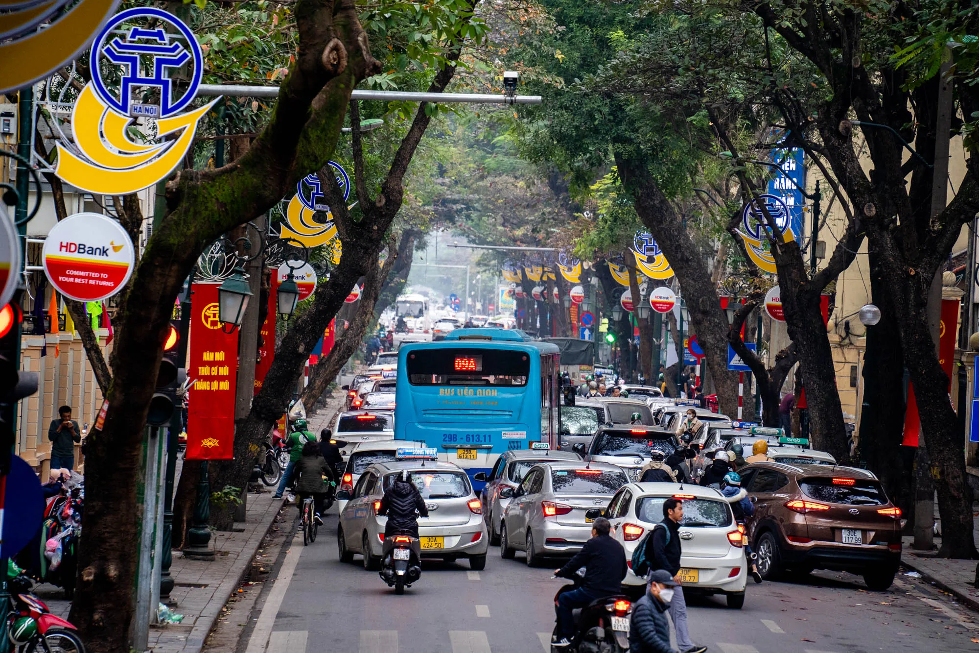 写真はベトナムのハノイで撮影された、混雑した通りの様子を捉えています。画面奥に向かって道路が伸び、車やバイクがひしめき合っています。中央には水色のバスが位置し、その周りを様々な車種の車が取り囲んでいます。多くの人がバイクに乗っており、歩行者も道路を横断しています。  道路の両脇には街路樹が植えられ、木々の間から空が見えます。街路灯には、ハノイのシンボルマークと思われる青と黄色の標識や、銀行の広告などが取り付けられています。赤い旗も飾られており、街に活気を与えています。全体的に曇り空で、少し霞んだような雰囲気が漂っています。交通量が多く、人々の喧騒が感じられる、ハノイの日常風景を切り取った一枚です。