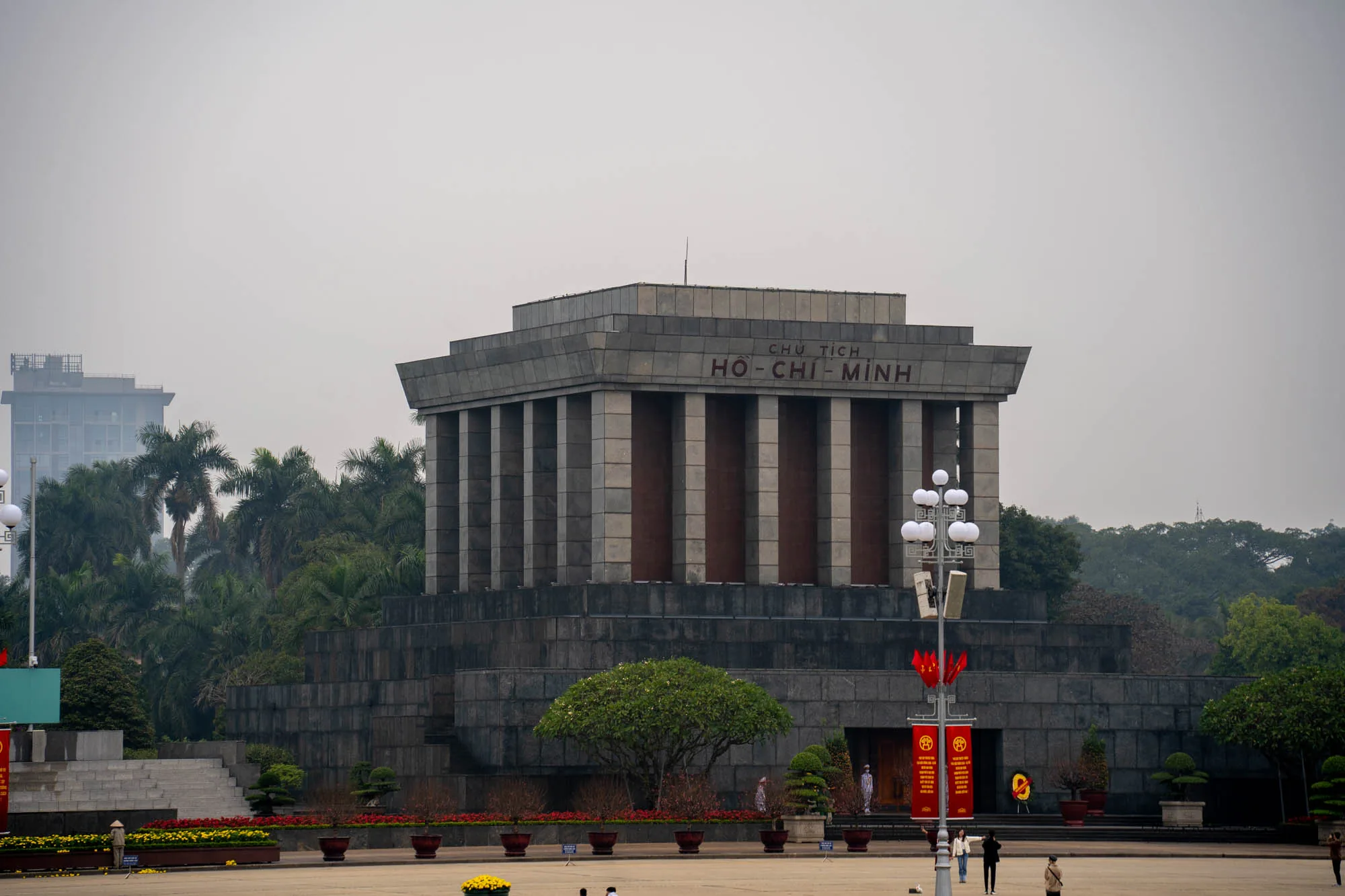 写真には、ベトナムのハノイにあるホーチミン廟が写っています。重厚な石造りの建物で、上部には「CHỦ TỊCH HỒ-CHÍ-MINH」と文字が刻まれています。建物の前には広場が広がり、人々がまばらに歩いています。広場には花壇や街灯があり、ベトナムの国旗を模した装飾が施されたポールが立っています。背景には緑豊かな木々が生い茂り、その奥には高層ビルが見えます。全体的に落ち着いた雰囲気で、少し曇り空のようです。厳粛で静謐な空気が漂っています。