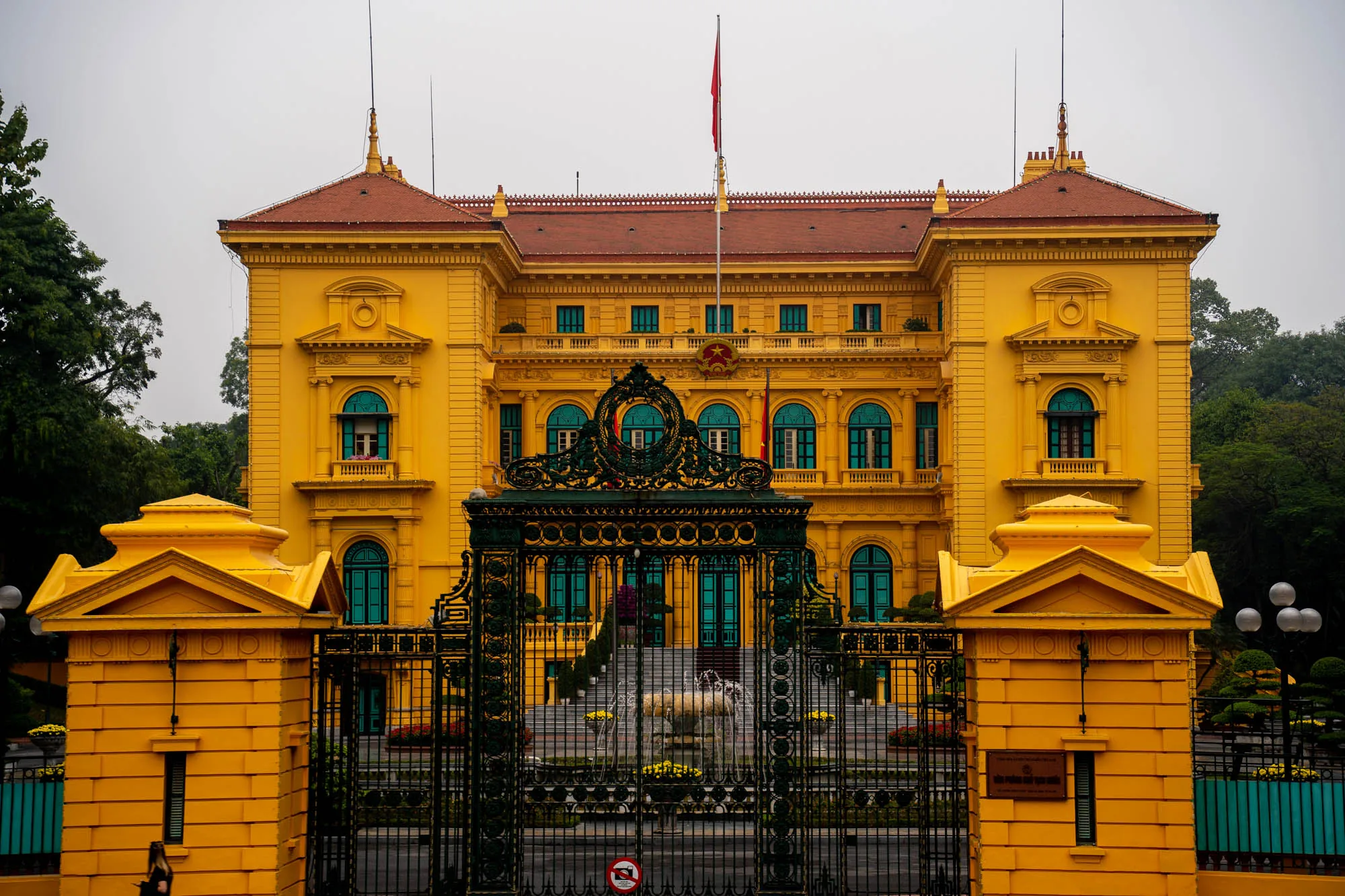 ベトナムのハノイで撮影された写真です。画面全体に、鮮やかな黄色の建物が大きく写っています。おそらく政府関連の建物だと思われます。建物の屋根は赤茶色で、窓枠やドアは鮮やかなターコイズブルーに塗られています。建物の正面には、装飾的な黒い鉄製の門があり、門の内側には噴水が見えます。建物の屋上にはベトナムの国旗が掲げられています。周囲には緑豊かな木々が生い茂り、全体的に少し曇った天気であることがうかがえます。建物の細部まで丁寧に描写されており、重厚感と歴史を感じさせる雰囲気です。