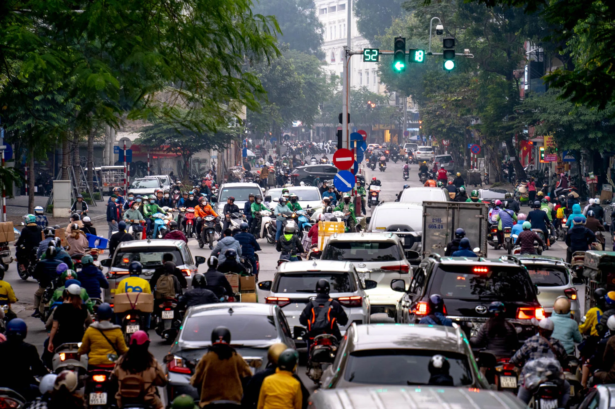 この写真は、ベトナムのハノイで撮影された交通風景を捉えています。道路は自動車とバイクで埋め尽くされており、かなりの交通渋滞が発生している様子が伺えます。手前には自動車が連なり、その後ろには無数のバイクがひしめき合っています。ほとんどのバイクの運転手はヘルメットを着用しており、様々な色の服を着ているのが見えます。多くの人が何かを運んでいるようです。背景には、緑豊かな街路樹と建物が立ち並び、交通信号機が設置されているのがわかります。信号機は青色を示しており、まだしばらくは交通の流れが続くことを示唆しています。全体的に、ハノイの活気ある、しかし混雑した日常の様子が伝わってきます。