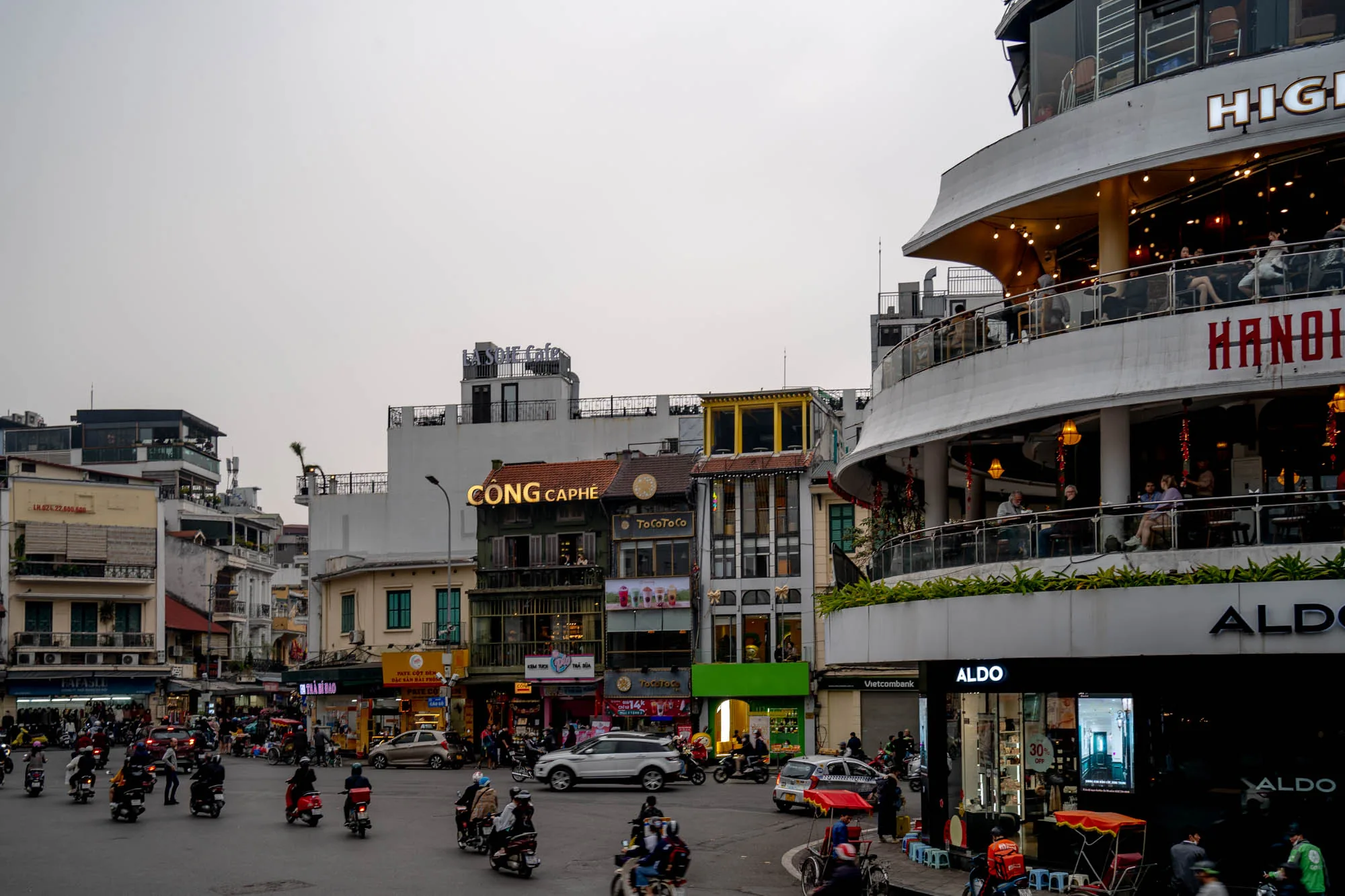 写真はベトナムのハノイで撮影された、活気あふれる街並みを捉えたものです。交差点には無数のバイクが走り、人々がそれぞれの目的地へと急いでいる様子が伺えます。空は曇り空で、全体的にやや霞んだ色調ですが、それがかえって街の喧騒を際立たせています。  道路沿いには様々な建物が立ち並び、カフェやショップが軒を連ねています。"CONG CAPHE"や"ToCoToCo"といった店名が看板に記されており、現地の雰囲気を醸し出しています。モダンな外観の建物には「HANOI」と大きく書かれ、複数の階に分かれたテラス席で人々が食事や会話を楽しんでいる様子が見えます。足元には靴のブランド「ALDO」の店舗もあり、現代的な商業の様子も伝わってきます。  交差点を行き交うバイクの数は非常に多く、交通量の多さが伺えます。歩道には物売りや客待ちの人がおり、生活感があふれています。全体的に、ハノイの活気とエネルギーを感じさせる一枚です。