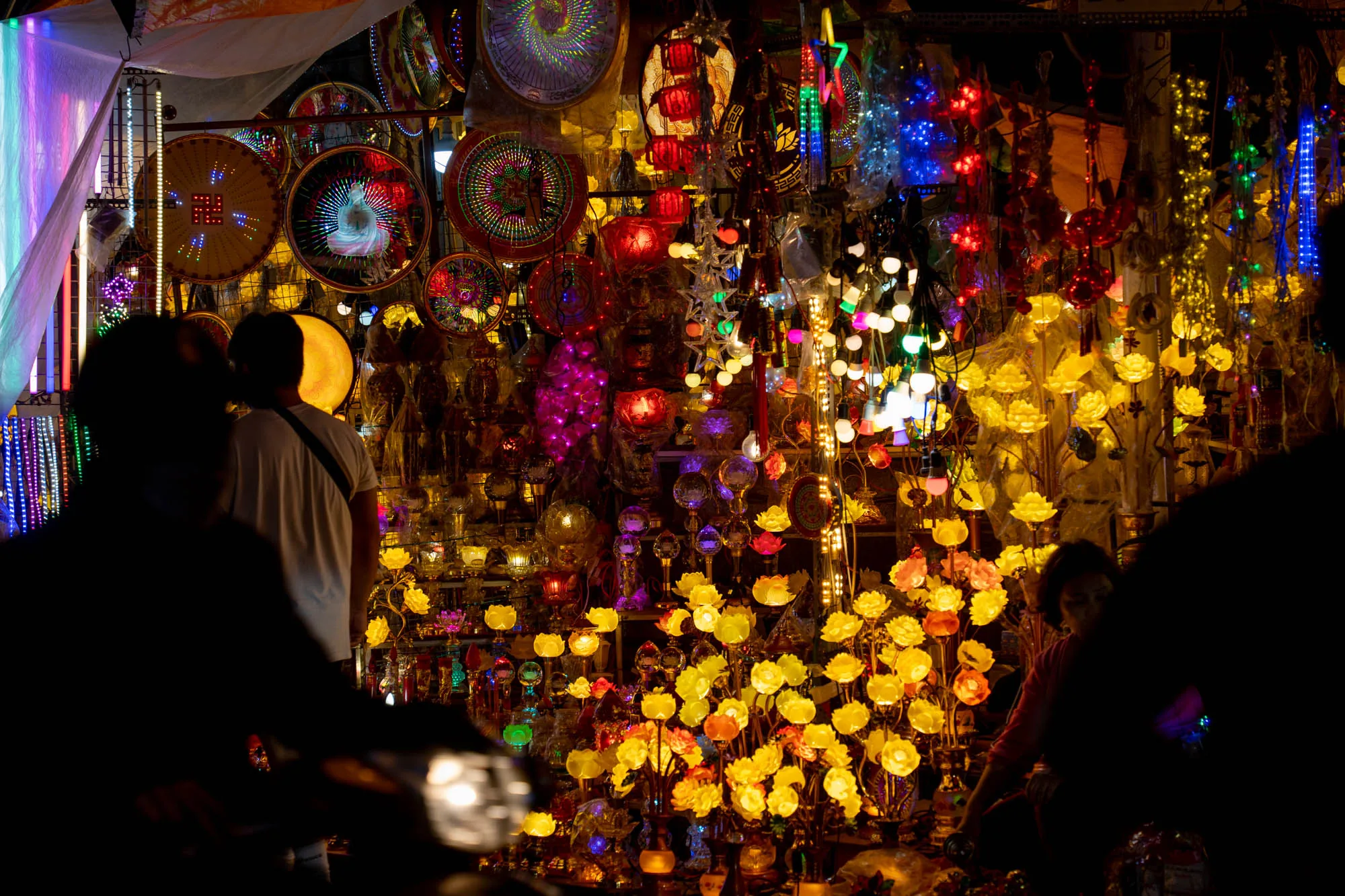 ベトナムのハノイで撮影された写真です。夜の市場と思われる場所で、色とりどりの照明や装飾品が所狭しと並べられています。特に目を引くのは、花を模した黄色やオレンジ色のライトで、温かい光を放っています。他にも、赤や青、紫など様々な色のライトが点滅し、賑やかで幻想的な雰囲気を醸し出しています。写真の手前には人のシルエットが見え、市場の活気を感じさせます。中央には、傘のような装飾品や、仏像が描かれた丸い装飾も見えます。全体的に暗い中で、様々な光が輝き、エキゾチックで独特な世界観を作り出しています。