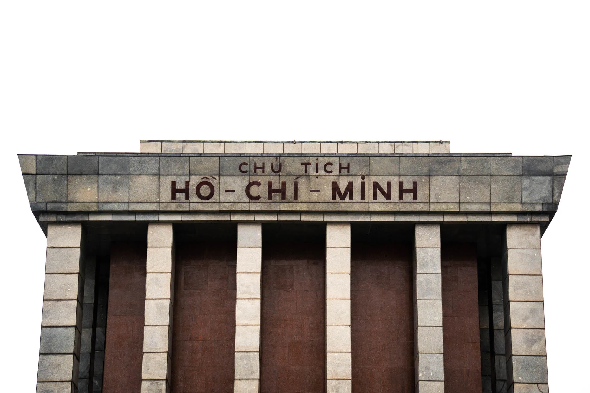 写真は、ベトナムのハノイにあるホー・チ・ミン廟の正面部分を捉えています。建物の外観は、グレーの石材で構成されており、重厚感と威厳を感じさせます。建物の上部には、赤褐色の文字で「CHỦ TỊCH HỒ CHÍ MINH」と書かれています。これは「ホー・チ・ミン主席」という意味です。下部には、赤褐色の壁面が並び、その間をグレーの石柱が規則的に配置されています。画面全体は白で切り取られており、建築物のディテールが際立っています。静かで厳粛な雰囲気が漂う写真です。