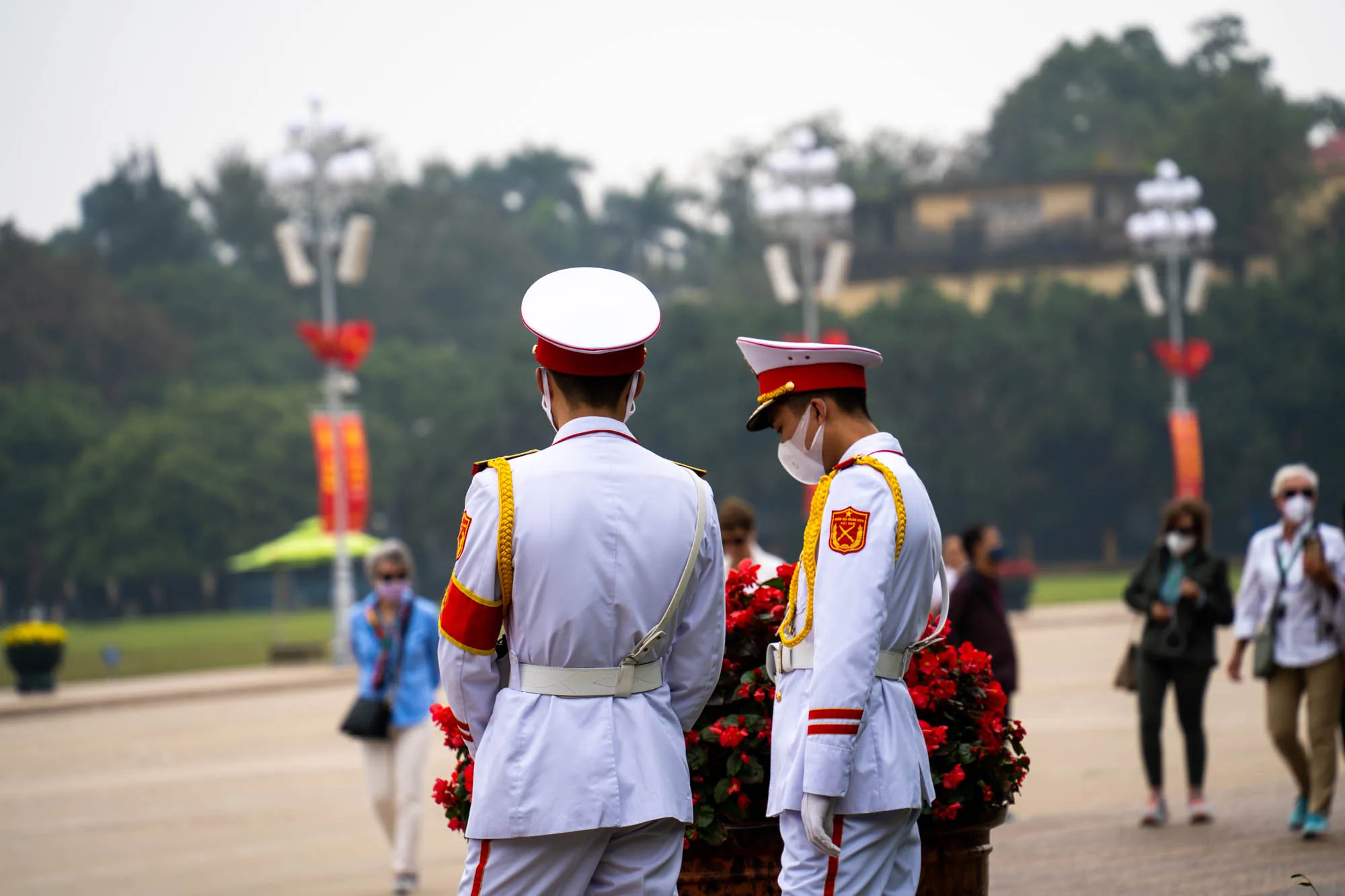 写真には、ベトナムのハノイで撮影されたと思われる、警備兵の姿が写っています。二人の警備兵は、白い制服に赤いラインが入った帽子をかぶっており、一人は白いマスクを着用しています。彼らは背を向けて立っており、手前には赤い花が植えられたプランターがあります。背景には、公園のような場所が広がっており、遠くに建物や木々が見えます。また、観光客のような人々が歩いている様子も確認できます。全体的に、静かで整然とした雰囲気の写真です。
