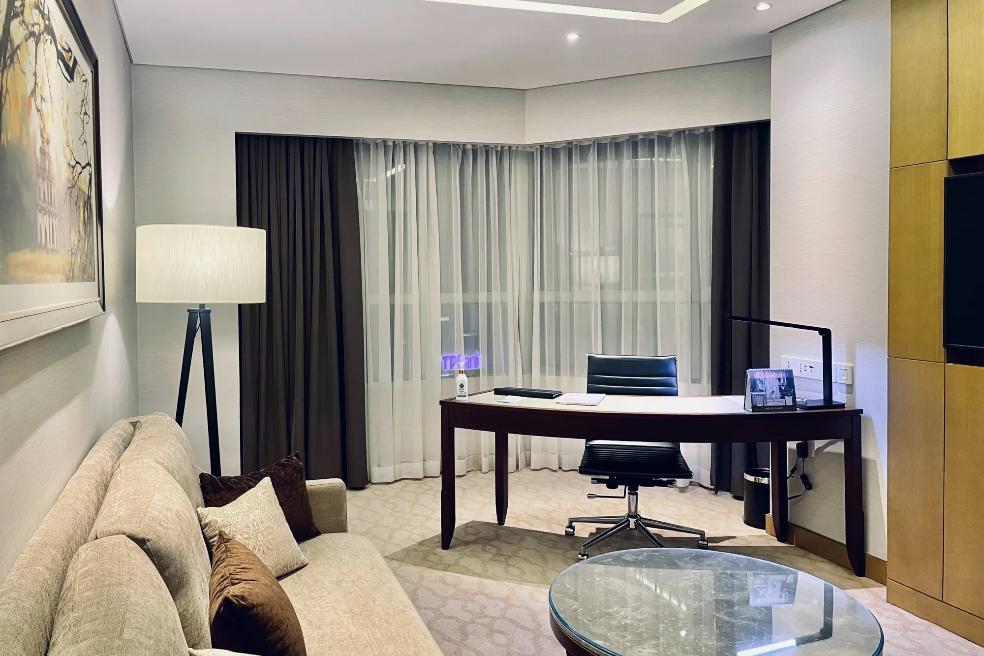 写真はベトナムのハノイで撮影されたホテルの部屋の様子です。室内は落ち着いた色調で統一されており、上品で洗練された雰囲気を醸し出しています。  部屋の左側にはベージュのソファが置かれ、茶色のクッションが置かれています。その隣には、黒い三脚スタンドのフロアランプが立っています。壁には絵画が飾られています。  部屋の中央には、木製のデスクと黒いオフィスチェアが配置されています。デスクの上には、書類、卓上カレンダー、消毒液、黒いデスクライトが置かれています。窓には茶色と白色のカーテンがかかっており、外の光を柔らかく遮っています。窓の外には街の灯りが見えます。  部屋の右側には、木製の収納棚とテレビが設置されています。手前には、ガラス製の丸テーブルが置かれています。  全体的に、快適で機能的な空間としてデザインされていることが分かります。ビジネスにも観光にも適したホテルの部屋であることが伺えます。