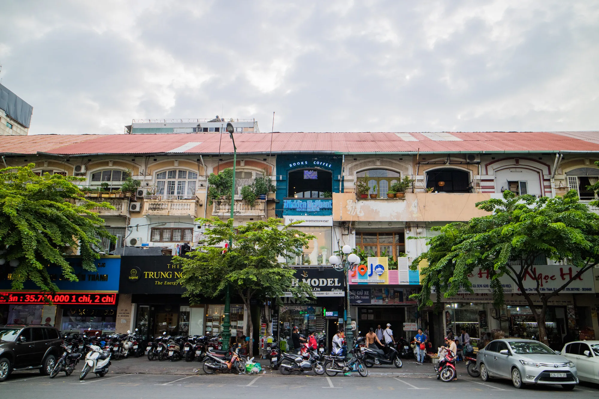 写真はベトナムのハノイで撮影されたもので、活気のある街の様子を捉えています。通り沿いには、赤茶色の屋根が特徴的な、古びた趣のある建物が連なっています。これらの建物は、それぞれ異なる色やデザインで装飾されており、多様な店舗や住居が入っているようです。上層階にはバルコニーが見え、植物が飾られているところもあります。  建物の一階部分には様々な店舗が並び、看板や広告がひしめき合っています。「BOOKS COFFEE」という店や、「MODEL」と書かれた看板が見えます。通りにはたくさんのバイクが駐輪されており、人々の往来も活発です。歩道で立ち話をする人や、バイクに乗る人、歩いている人が写っており、ハノイの日常的な風景が伝わってきます。  空は曇っており、落ち着いた雰囲気ですが、街全体はエネルギーに満ち溢れているように感じられます。緑豊かな木々が建物の間に植えられており、都市景観に自然の要素を加えています。写真全体を通して、ハノイの独特な魅力と、アジアの都市ならではの喧騒が感じられる一枚です。