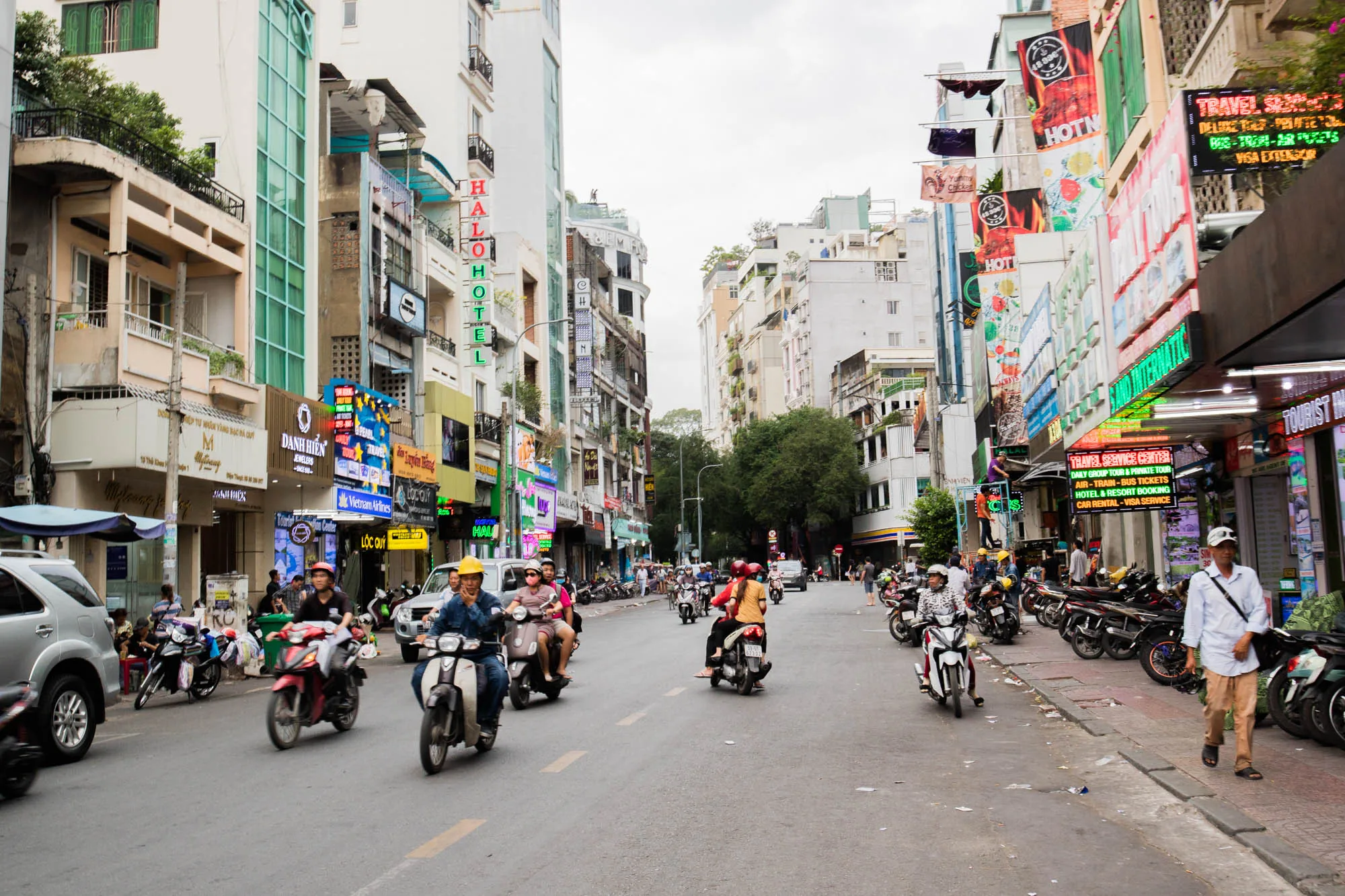 写真はベトナムのハノイの通りを捉えたもので、日常の喧騒が感じられます。画面中央には、多数のバイクが道路を走行しており、ハノイの主要な交通手段であることが伺えます。歩道にはバイクが駐輪され、歩行者が歩いています。建物の壁面には、ネオンサインやカラフルな看板が所狭しと並び、現地の活気ある商業活動を物語っています。看板には「HALO HOTEL」「Vietnam Airlines」などの文字が見え、ホテルや航空会社の広告であることがわかります。また、「TRAVEL SERVICE CENTER」と書かれた看板もあり、観光客向けのサービスを提供している場所のようです。空は曇り空で、全体的に落ち着いた光のトーンですが、ネオンサインの光が街を明るく照らし出しています。写真全体から、東南アジア特有のエネルギーと活力が伝わってきます。