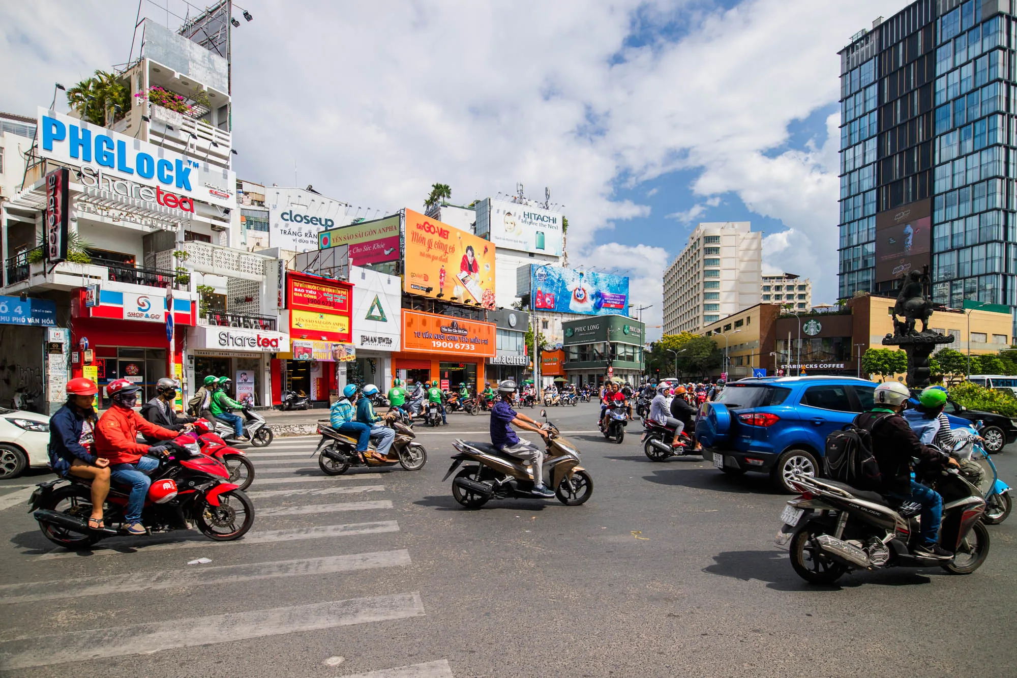 この写真はベトナムのハノイで撮影された、非常に活気のある都市の風景を捉えています。画面全体にバイクがひしめき合い、交差点を行き交う様子が写されています。多くの人がヘルメットを着用しており、中には二人乗りをしている人も見られます。道路はアスファルトで舗装されており、白い横断歩道が引かれています。  背景には、様々な高さの建物が立ち並び、特に目を引くのは、複数の大きな広告看板です。看板には、PHGLock、Sharetea、Hitachi、Phuc Longなどのブランド名が確認できます。また、看板の多くはベトナム語で書かれており、現地の雰囲気を伝えています。  写真の右側には、ガラス張りの高層ビルと、スターバックスの店舗が見えます。これらの建物は、ハノイの近代化された側面を象徴しているようです。空は晴れており、白い雲が浮かんでいます。全体的に、写真からはハノイのエネルギーと活気が感じられます。交通量が多く、看板や建物が密集していることから、都市部の混雑した様子が伺えます。