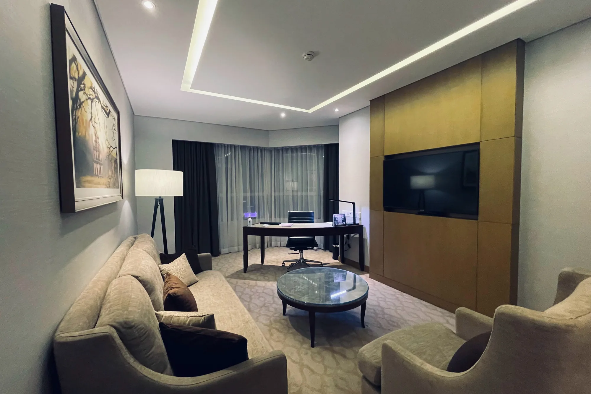この写真は、ベトナムのハノイにあるホテルの客室を捉えたものです。室内はベージュとブラウンを基調とした落ち着いた色調で統一されており、上品で洗練された雰囲気を醸し出しています。  部屋の中央には、ガラス天板の丸いコーヒーテーブルが置かれ、その周囲にはベージュのソファとアームチェアが配置されています。ソファには複数のクッションが置かれ、快適な空間を演出しています。  部屋の奥には、仕事用のデスクと椅子が設置されています。デスクの上には、書類や照明器具が置かれ、機能的なスペースとなっています。窓には薄手のカーテンと遮光カーテンが掛けられており、日差しの調節が可能です。  壁際には、大きな絵画が飾られ、部屋の雰囲気を高めています。また、壁には大型のテレビが埋め込まれており、エンターテイメントを楽しむことができます。天井には間接照明が施されており、部屋全体を優しく照らしています。全体的に、この部屋は快適性と機能性を兼ね備えた、くつろげる空間となっています。