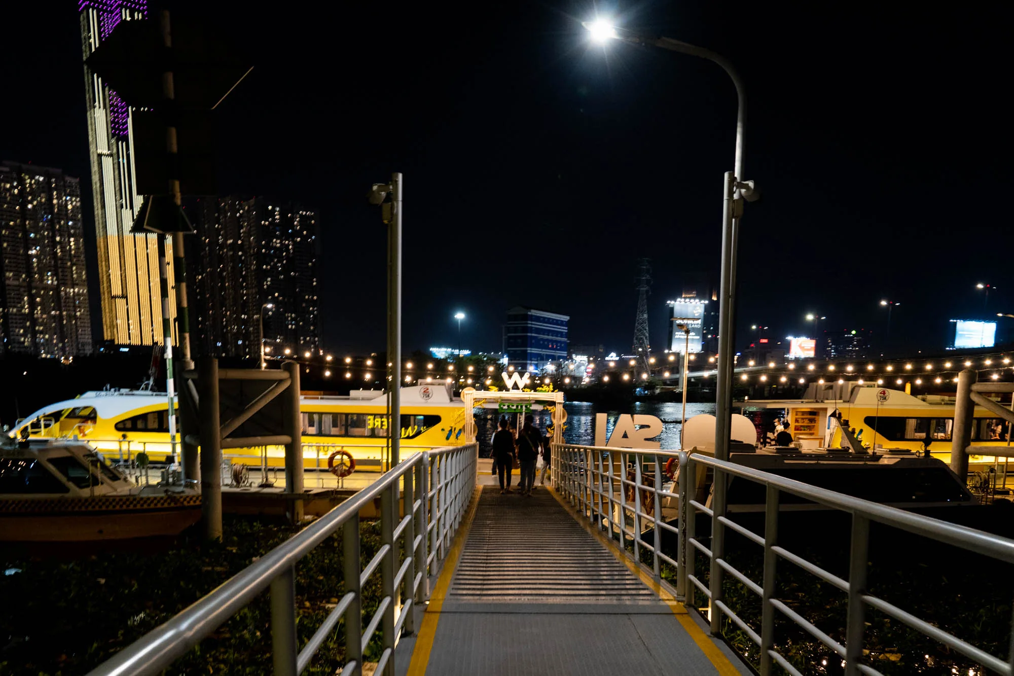 写真はベトナムのホーチミンで撮影された夜景です。手前には金属製の柵に囲まれた桟橋が写っており、数人の人物がその上を歩いています。桟橋の先には、黄色の船体が特徴的な観光船が複数停泊しています。背景には、高層ビル群がそびえ立ち、ライトアップされた建物やネオンサインが夜空を彩っています。街灯が桟橋を照らし、水面には光が反射してきらめき、幻想的な雰囲気を醸し出しています。ホーチミンの賑やかな夜の様子が伝わってくる一枚です。